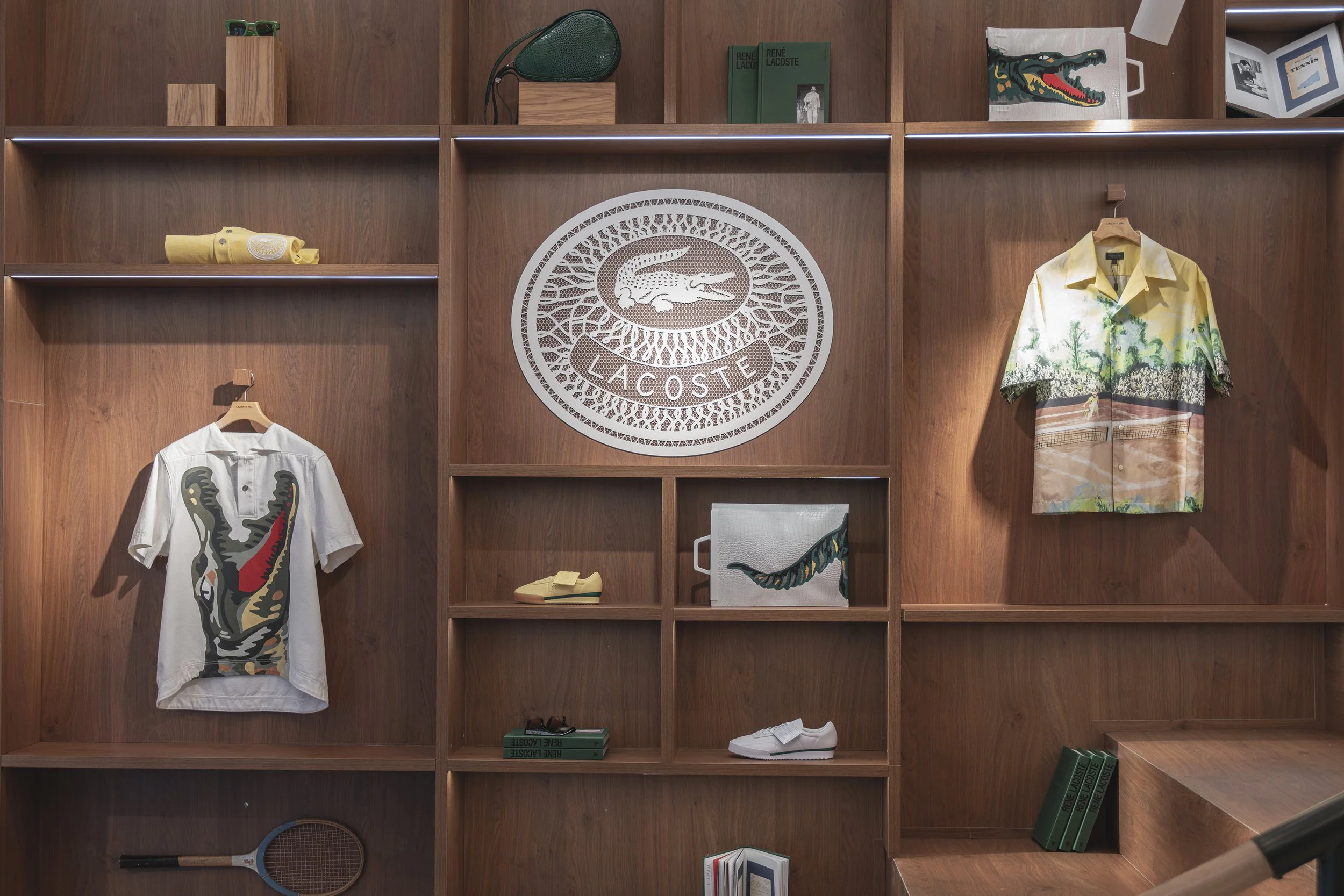 Étagères en bois avec des vêtements, sacs et livres de la marque Lacoste, dont deux chemises avec motifs de crocodile et une grande affiche du logo Lacoste.