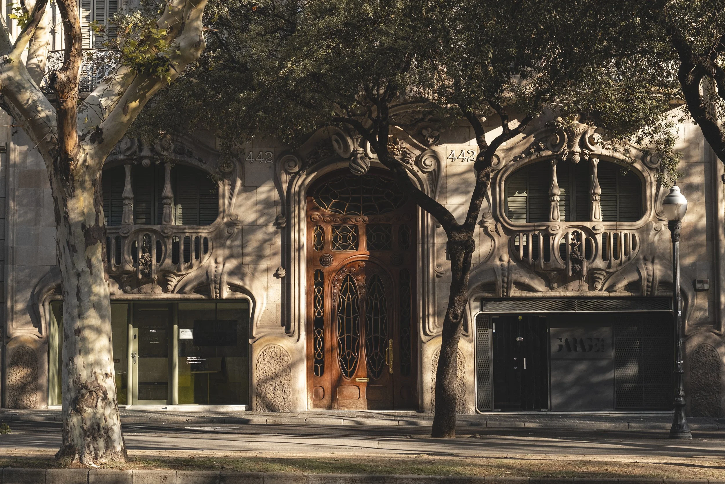 Bâtiment artistique avec façade en stuc orné de détails sculptés, fenêtres ovales avec balustrades en pierre et porte en bois au centre, entouré d'arbres et éclairé par la lumière du soir.