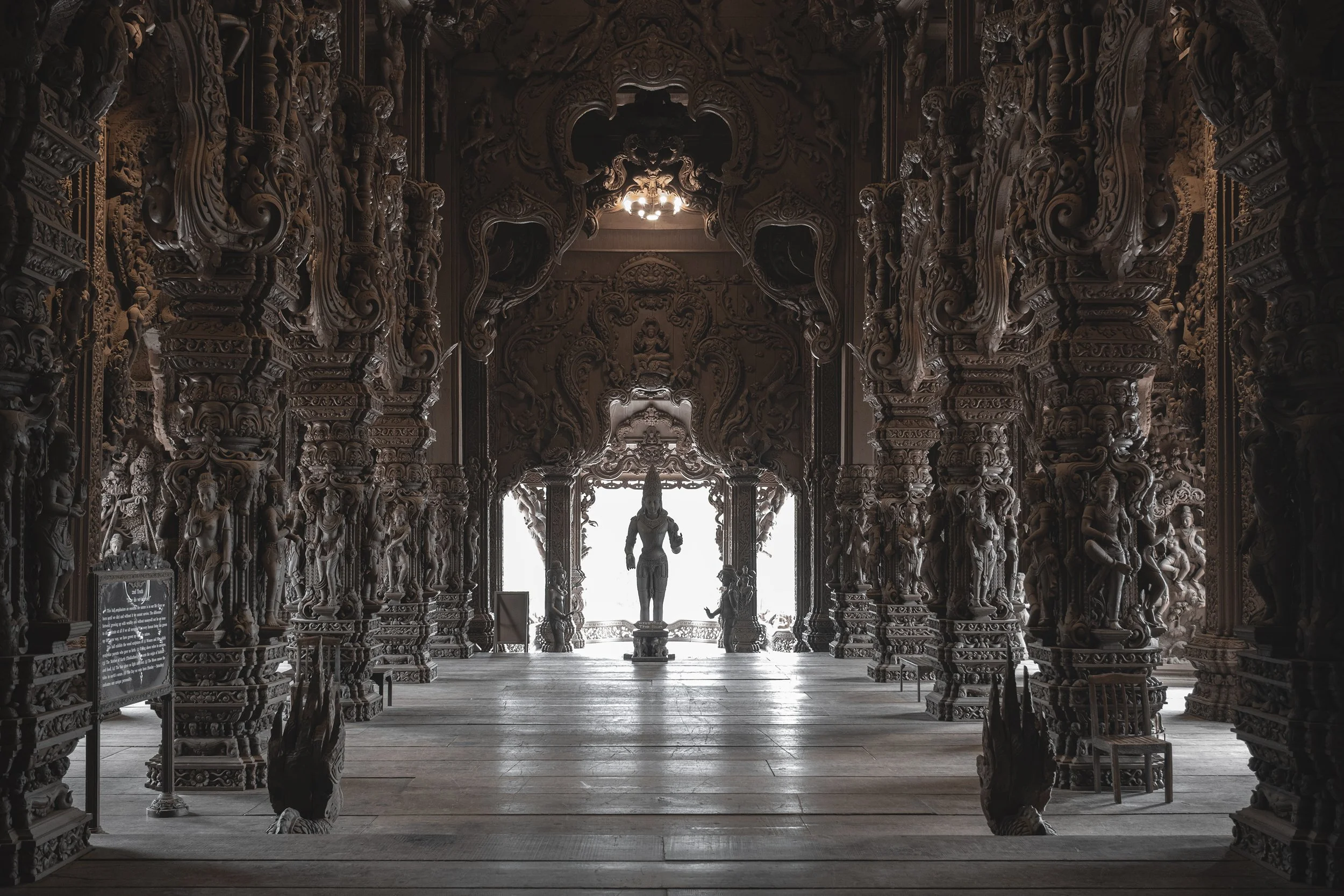 Intérieur d'un temple en bois sculpté avec de nombreuses statues et détails artistiques, lumière venant par l'arrière, vue vers une statue centrale en arrière-plan.