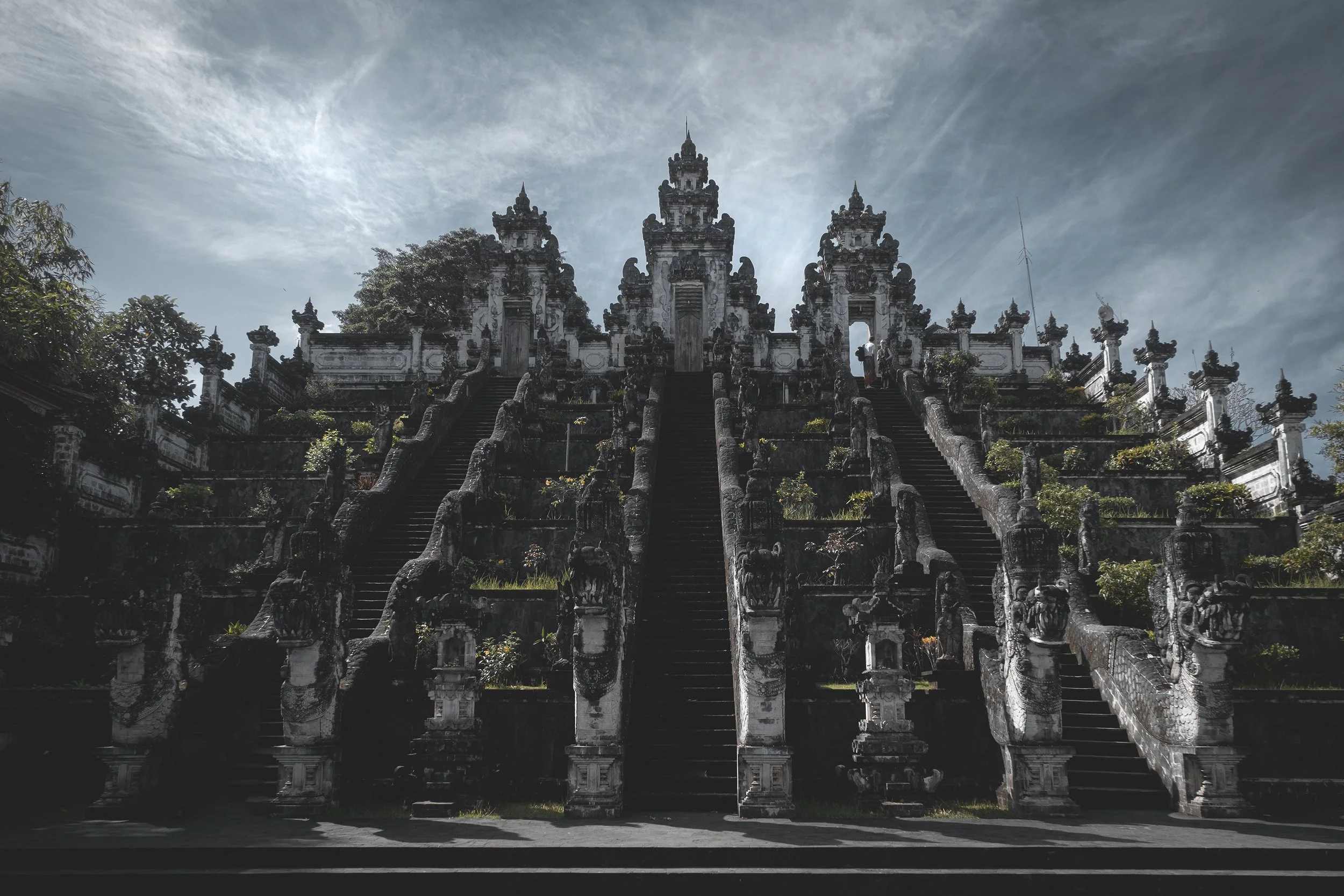 Un temple ancien avec plusieurs escaliers menant à des portes, décoré de statues et de végétation, sous un ciel nuageux.