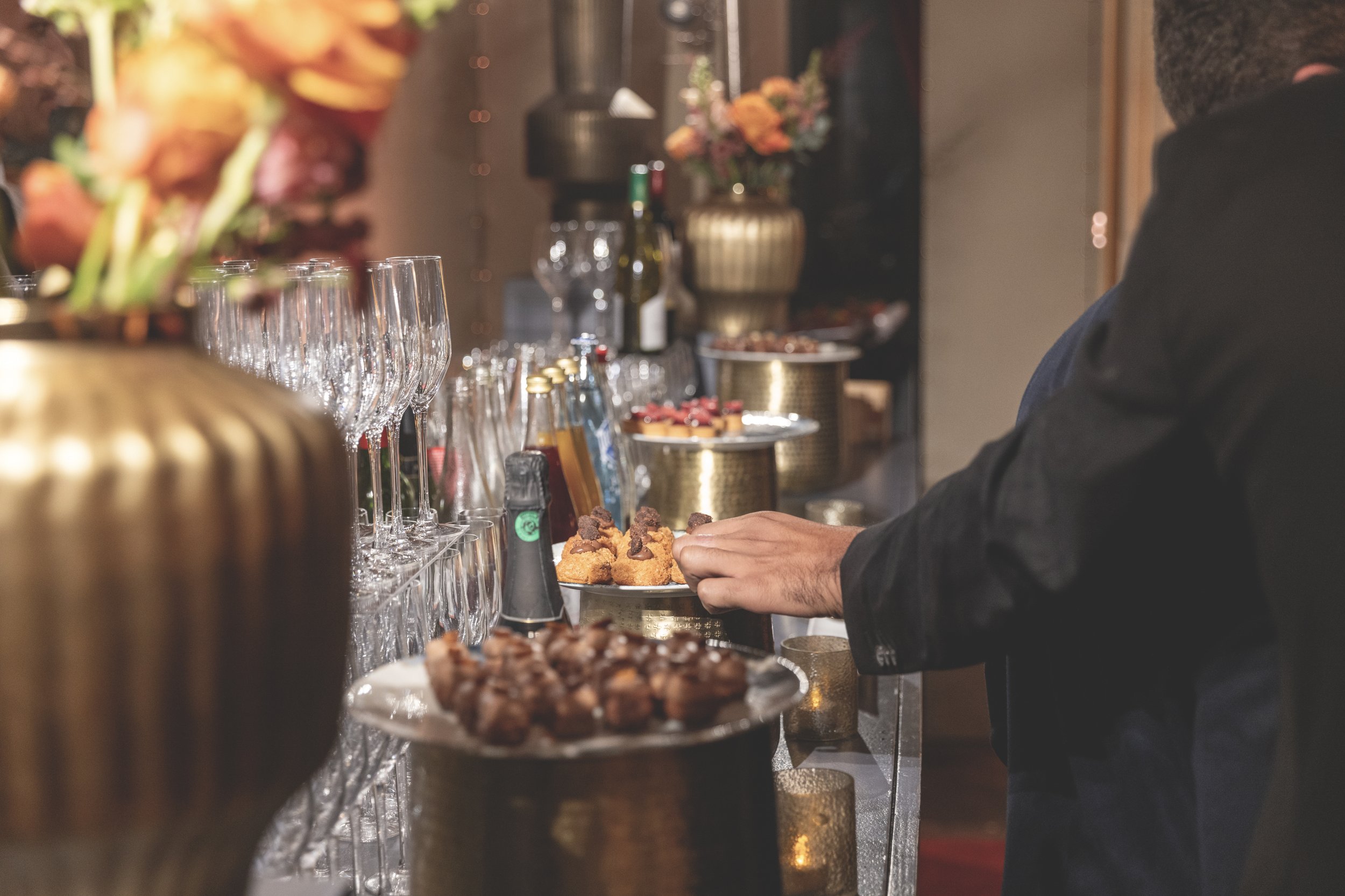 Une personne sert des amuse-gueules lors d'une réception ou d'un événement, avec des verres à vin et des bouteilles sur une table élégante.