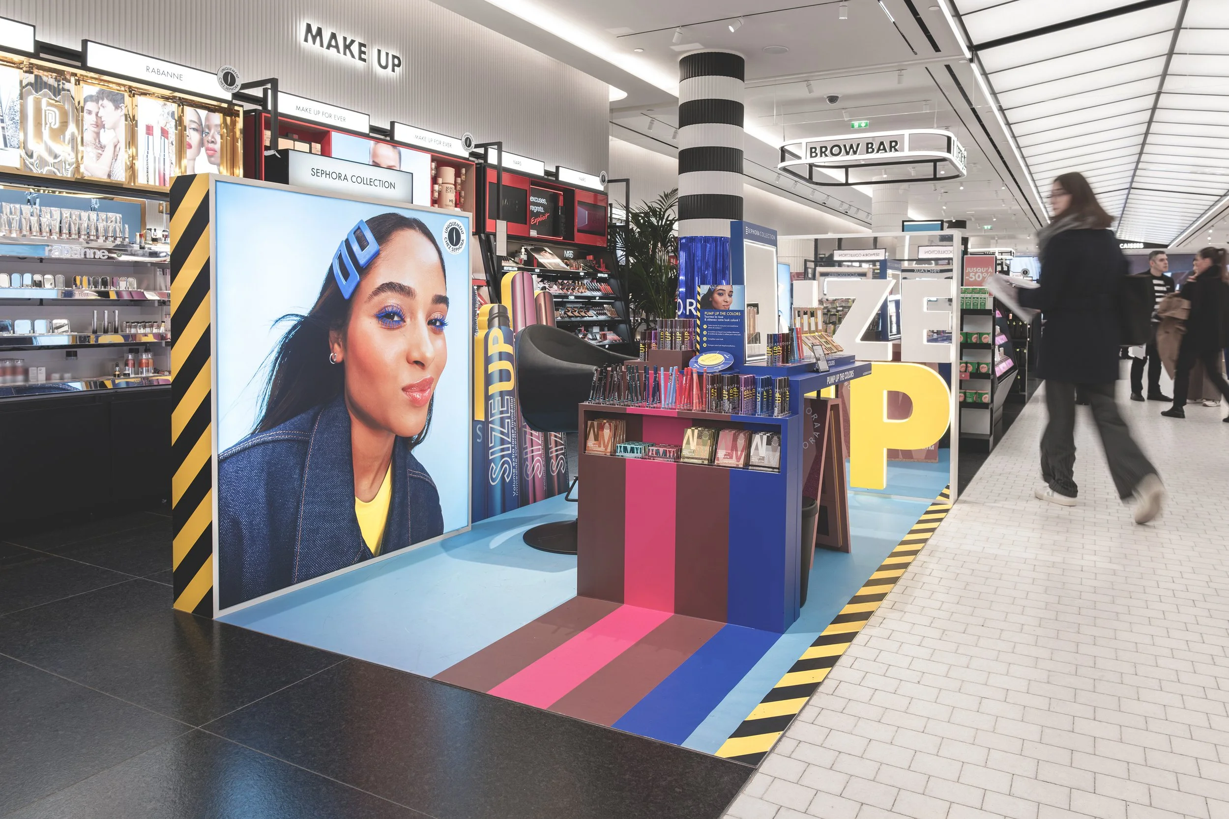 Zone de maquillage et produits de beauté dans un centre commercial, avec une grande affiche promotionnelle d'une femme portant un serre-tête bleu, des stands colorés et des rayons avec différentes marques cosmétiques.