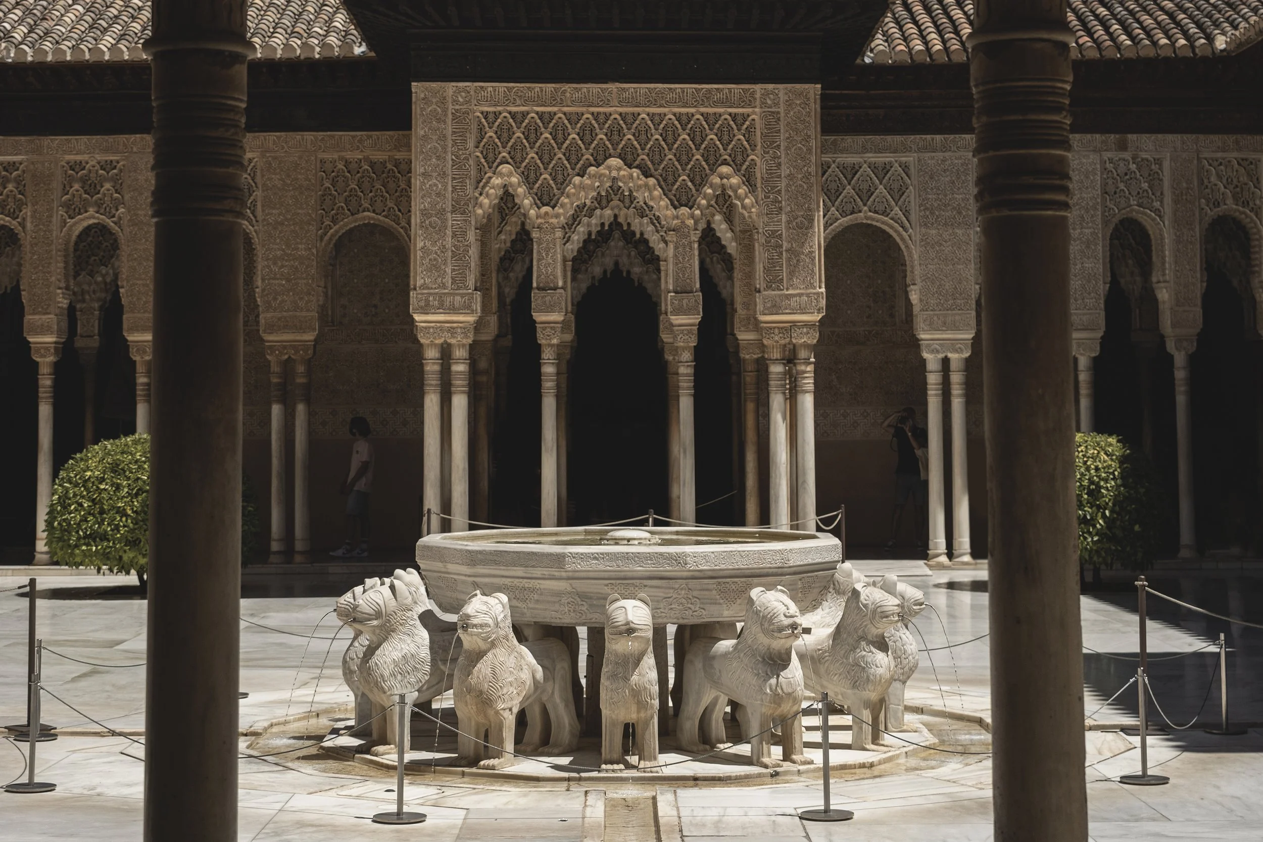 Un cour intérieur d'un bâtiment avec une fontaine ornée de lions en marbre, entourée de colonnes et d'arcs décorés dans un style islamique, avec deux personnes visibles à l'arrière.