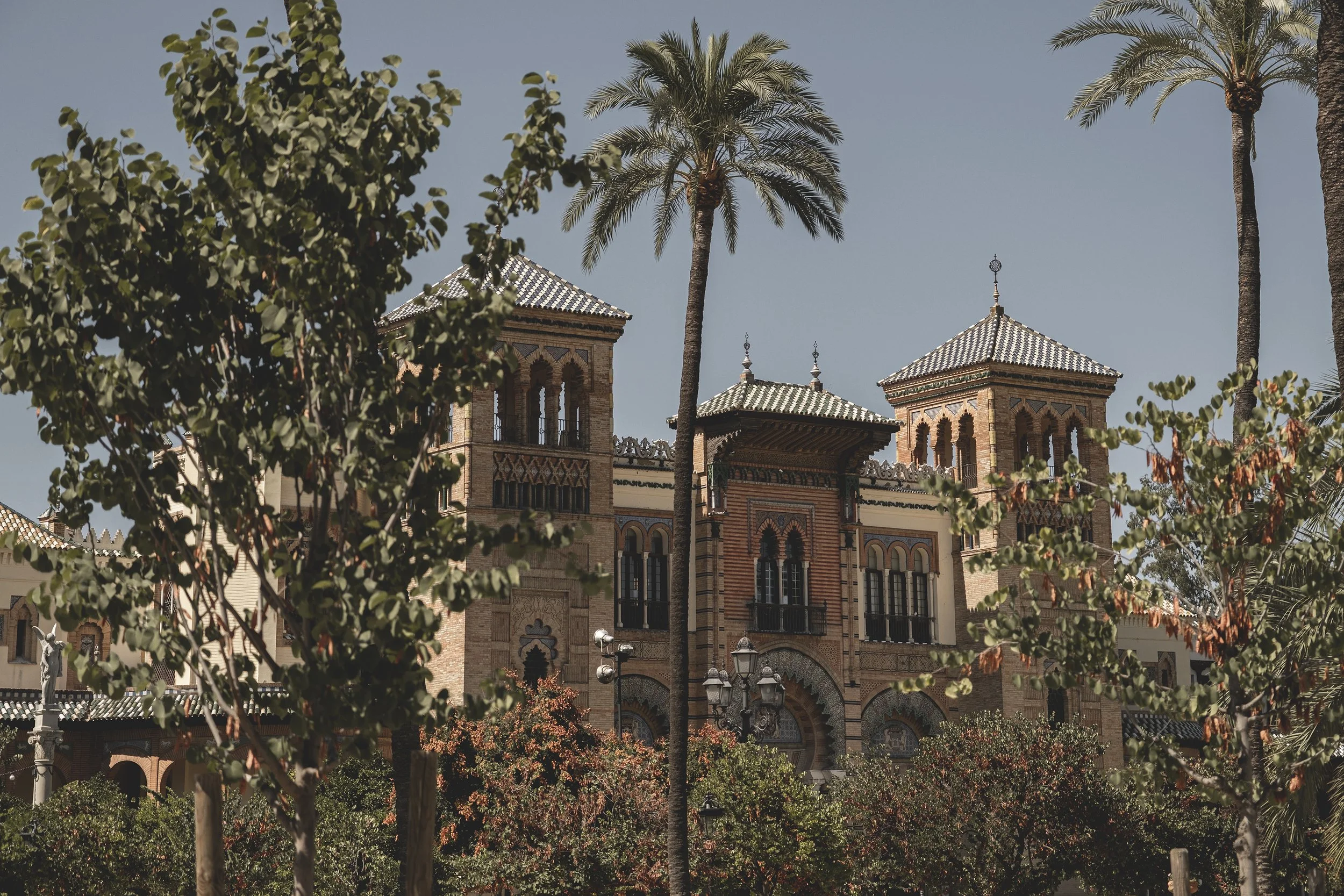 Une vieille bâtisse en pierre avec des tours et des fenêtres arqué, entourée de palmiers et d'arbres, sous un ciel bleu clair.