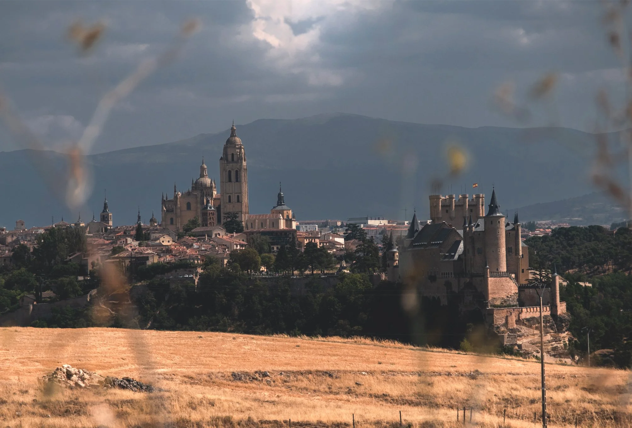 Vue d'un château et une église sur une colline, avec des montagnes en arrière-plan, vue à travers des branches floues en premier plan, sous un ciel nuageux.