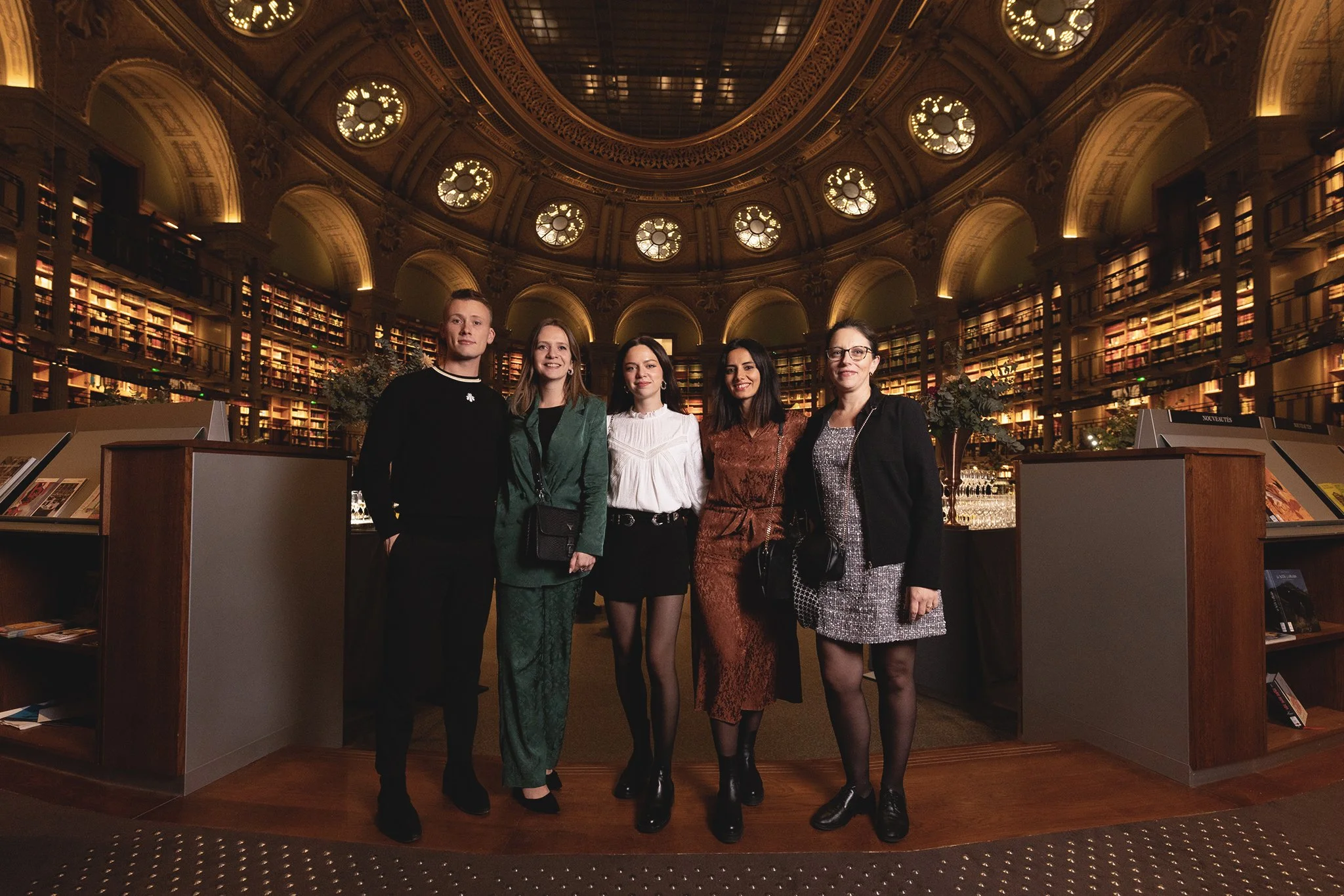 Cinq femmes posant dans une bibliothèque somptueuse avec de hautes étagères remplies de livres, un dôme en verre avec des lumières circulaires au plafond