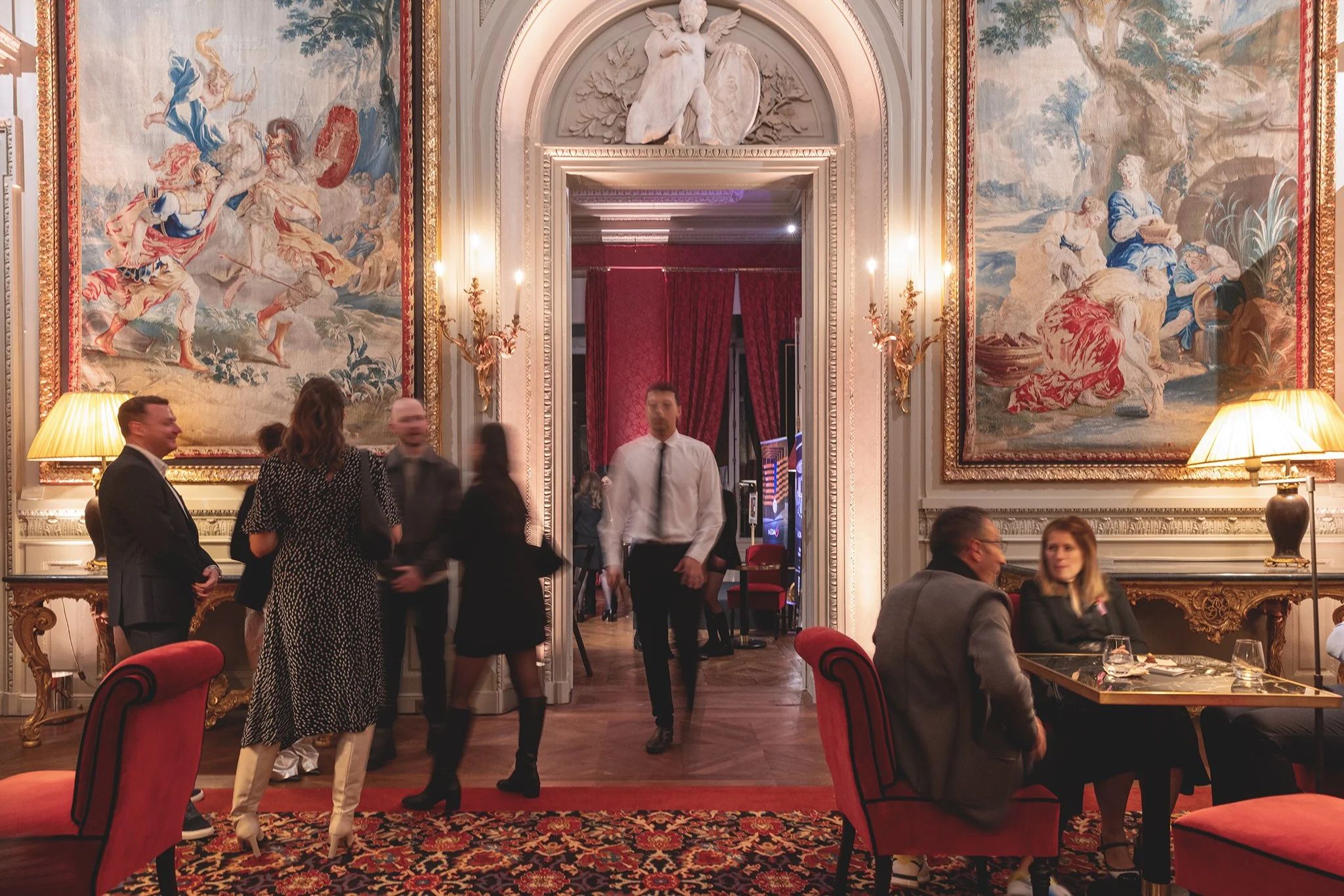 Intérieur d'un restaurant élégant avec des murs ornés de grands tapisseries, des lampes à abat-jour, des tables et des chaises rouges, et des personnes en tenue de soirée discutant et dégustant leur repas.