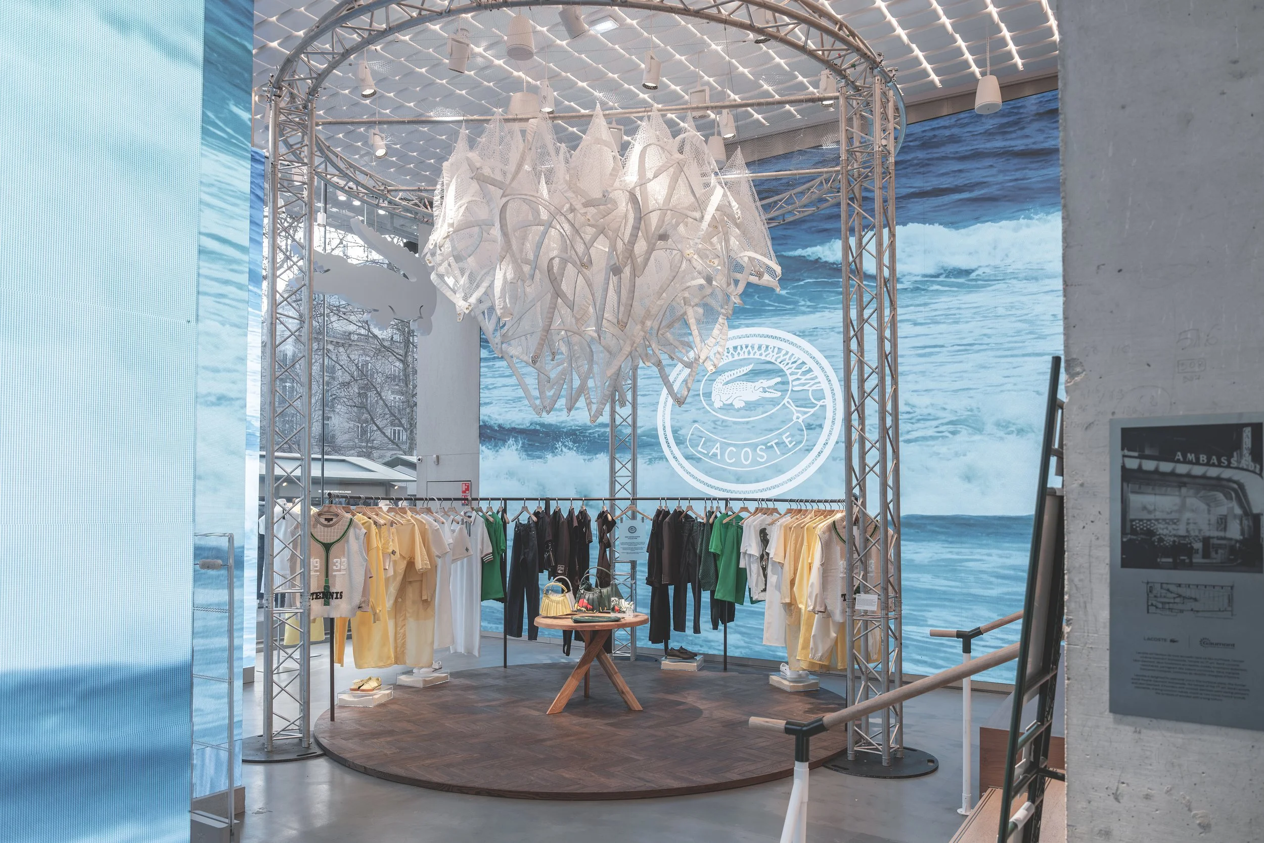 Espace de vente de vêtements Lacoste, avec des chemises, t-shirts et pantalons de différentes couleurs, sous un éclairage moderne, avec un grand logo Lacoste en arrière-plan représentant un crocodile dans un décor nautique.