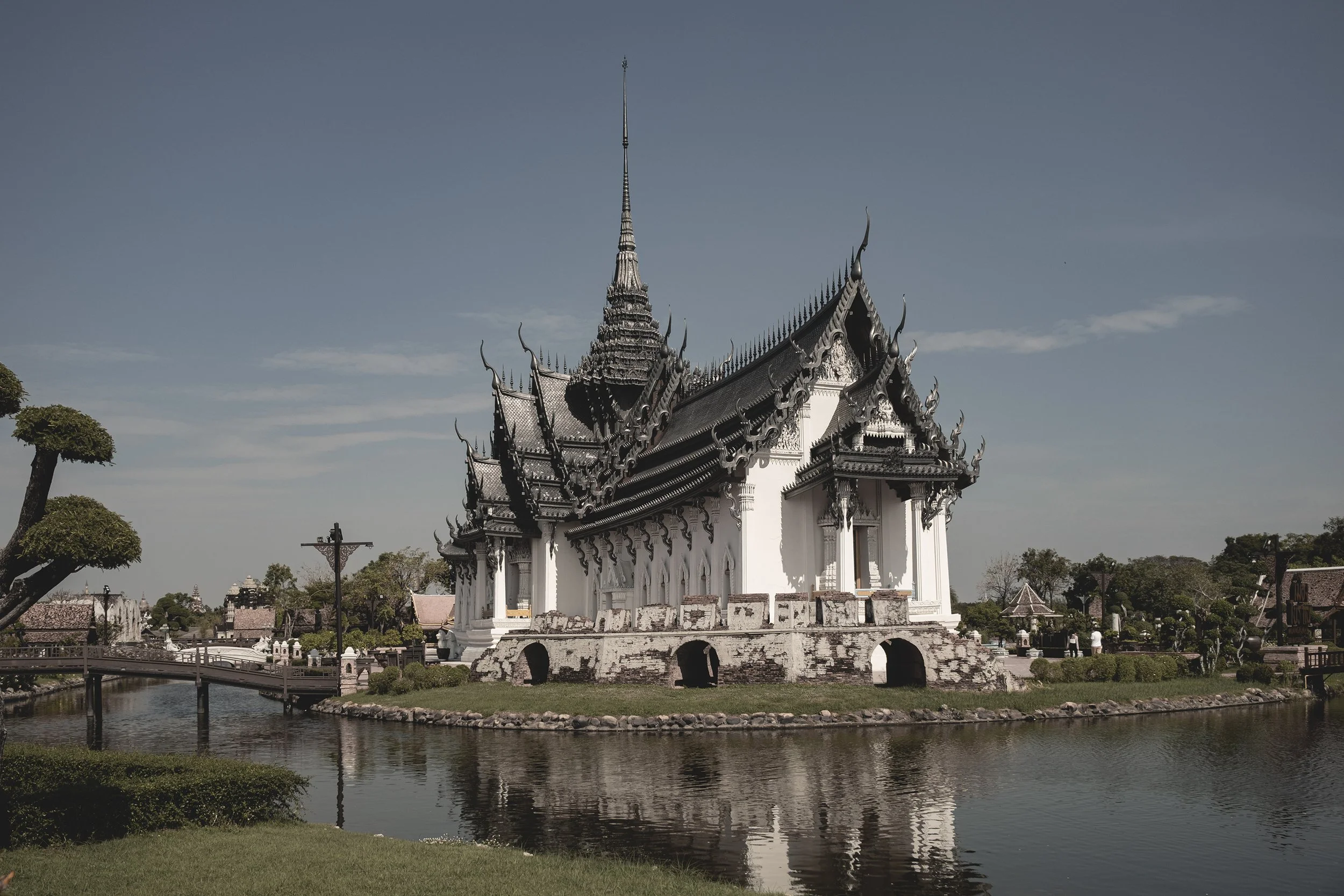 Un temple traditionnel thaïlandais blanc avec un toit en pente orné de sculptures en forme de serpents, entouré d'eau et de jardins verdoyants.