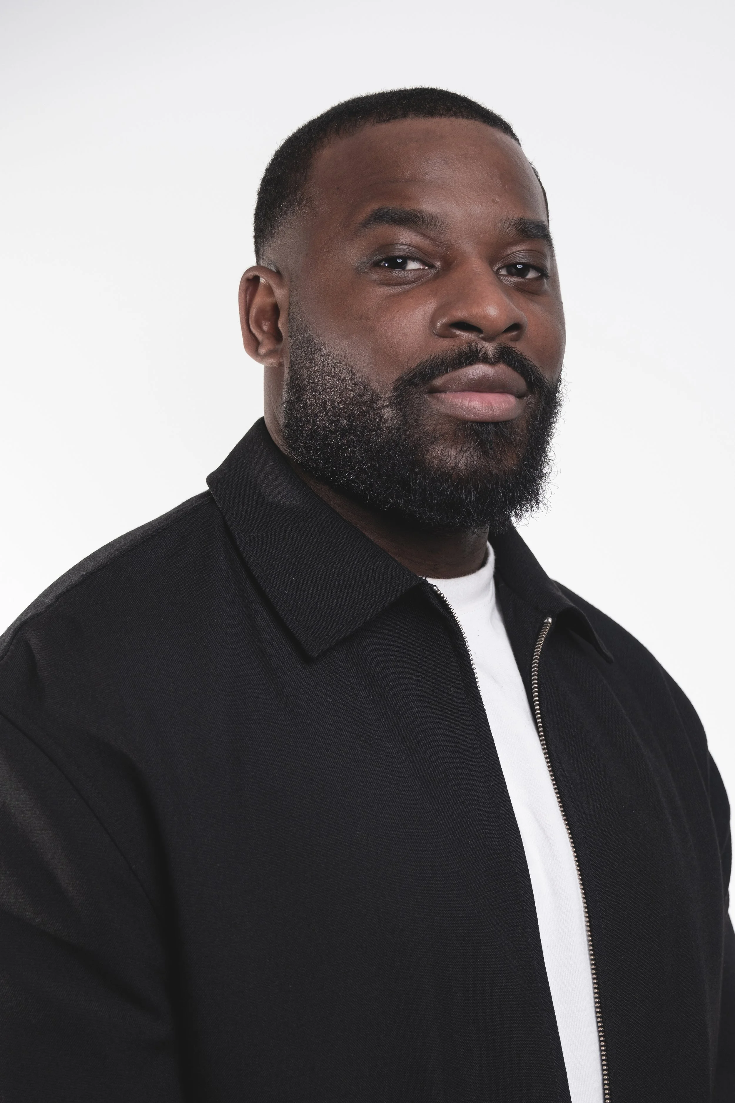 Portrait d'un homme noir avec une barbe, portant une veste noire et une chemise blanche contre un fond blanc.
