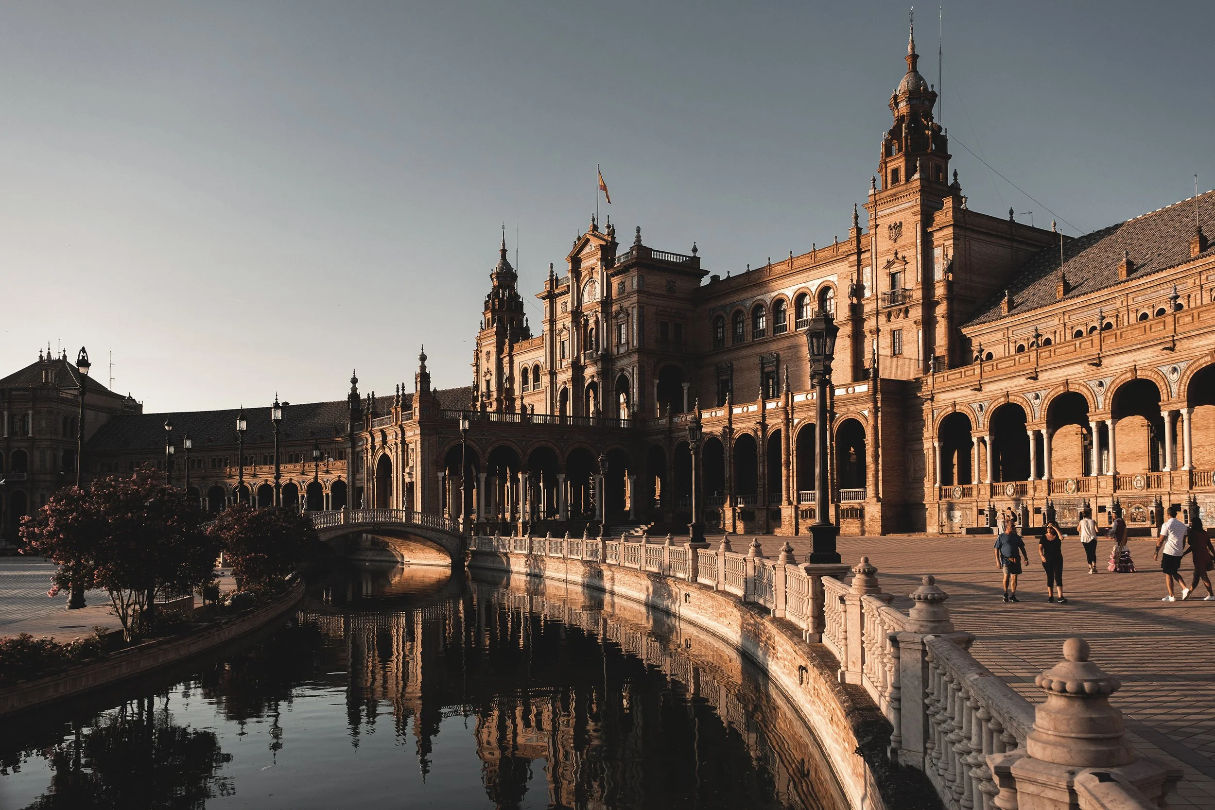 Photo d'un bâtiment historique avec une grande façade en pierre, entouré d'une promenade en arc de cercle et d'une petite rivière refletant le bâtiment, sous un ciel clair et ensoleillé, with des personnes se promenant.