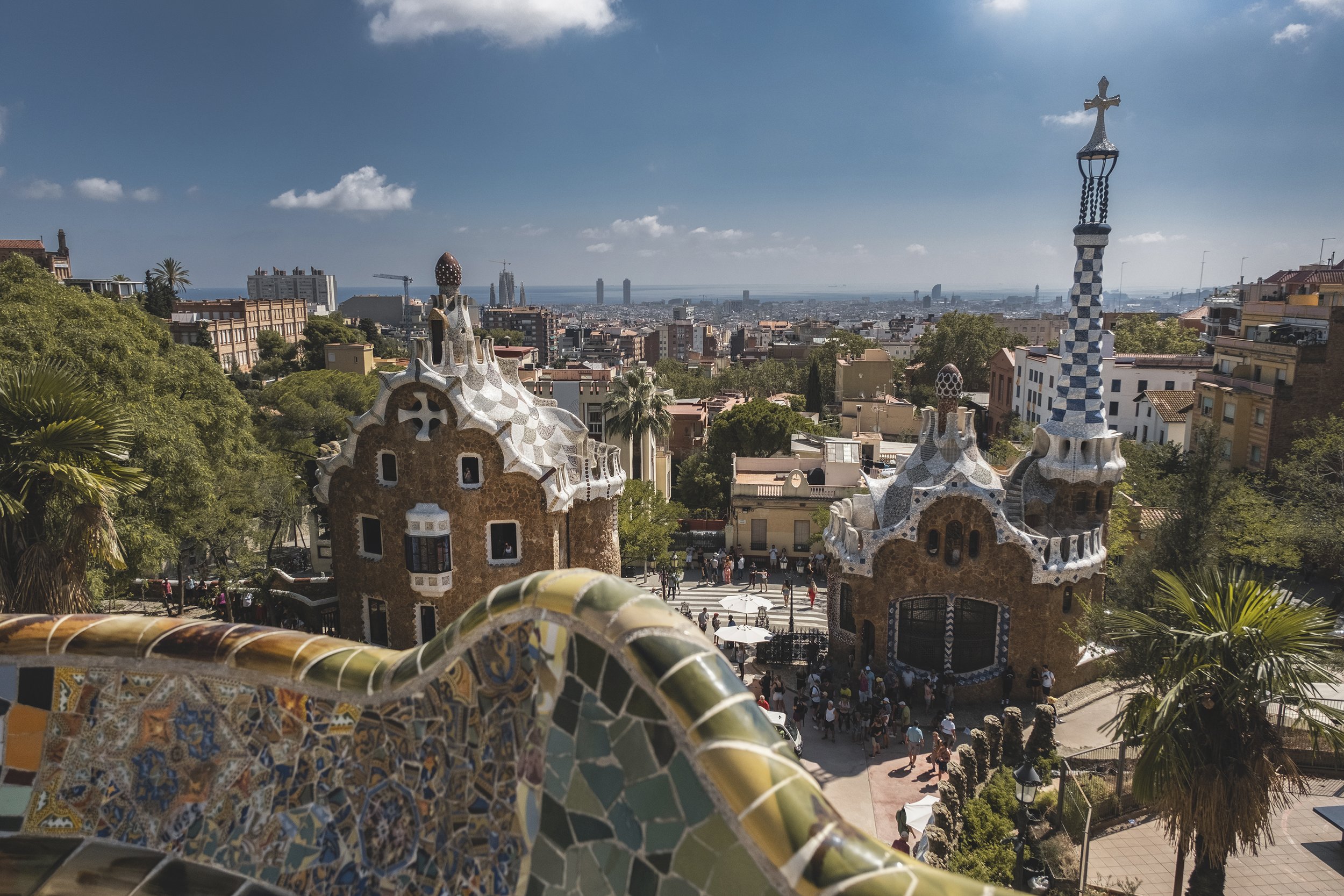 Vue de l'architecture colorée et curviligne de la Parc Güell à Barcelone, avec des bâtiments en mosaïque, arbres, personnes en bas, et la ville au fond sous un ciel bleu.