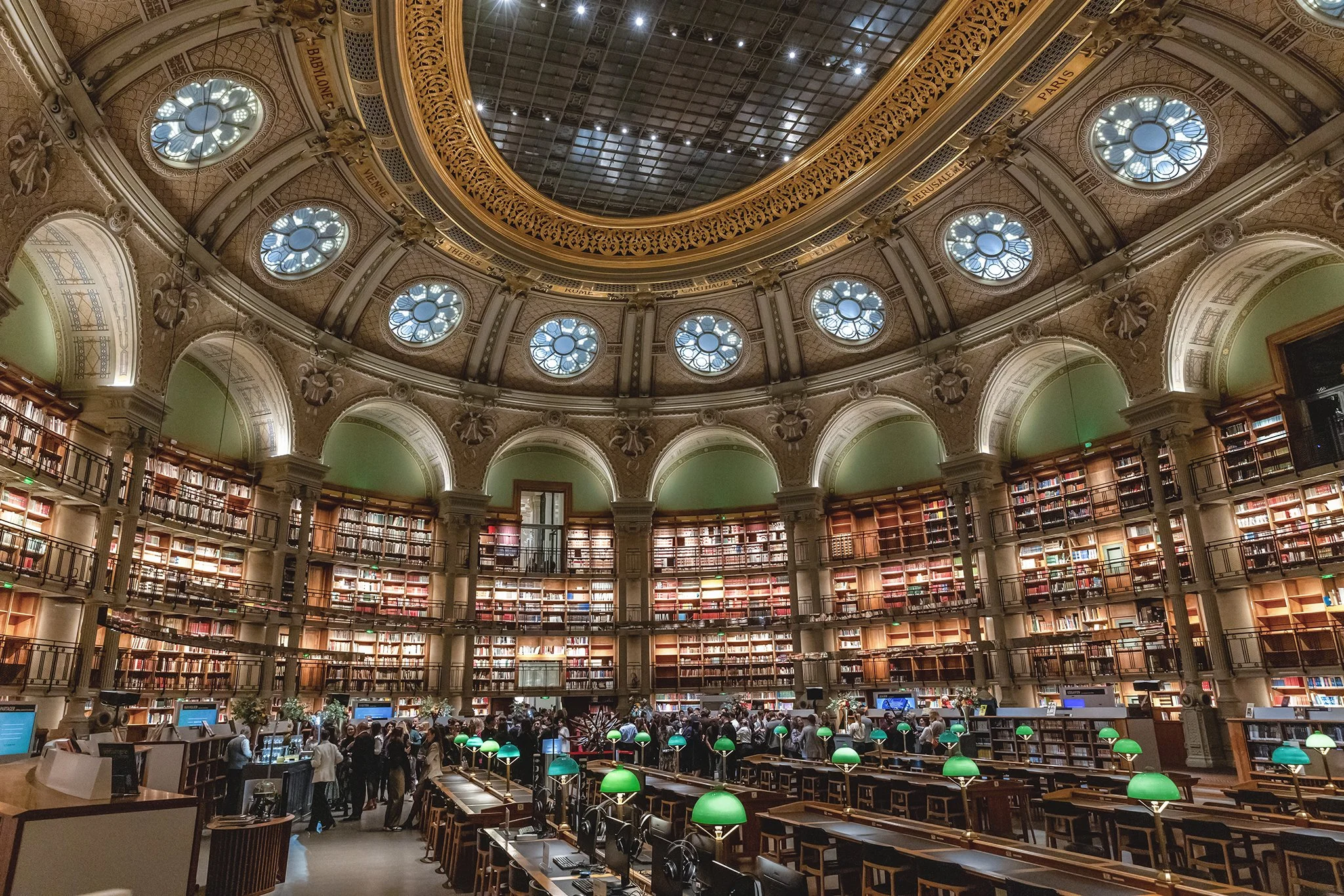 Une grande bibliothèque historique avec une coupole en verre, des étages remplis de livres, et des personnes utilisant des ordinateurs et lisant des livres.
