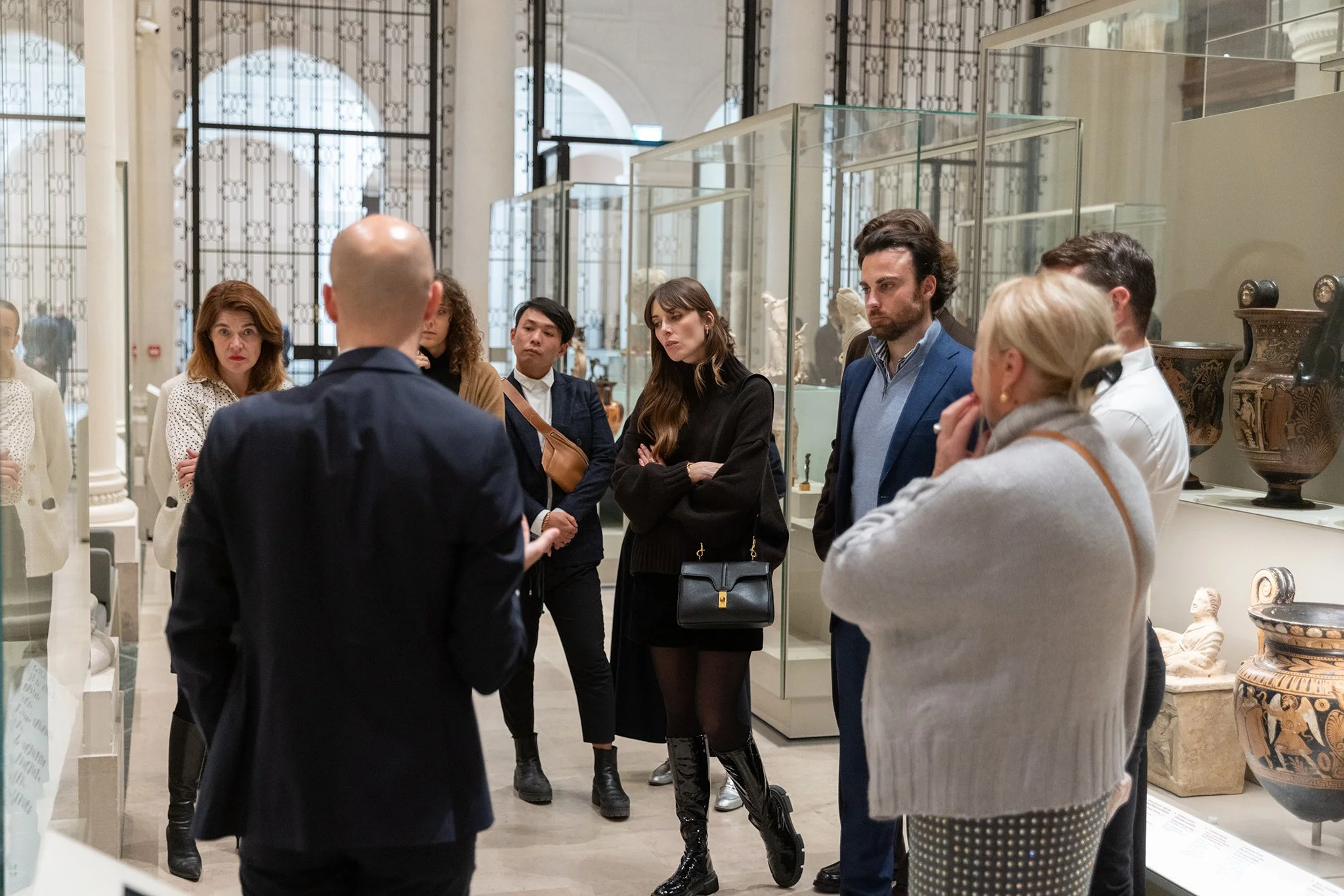 Groupe de personnes dans un musée en train d'écouter un guide ou un conférencier, avec des vitrines et des objets d'art ou antiques en arrière-plan.