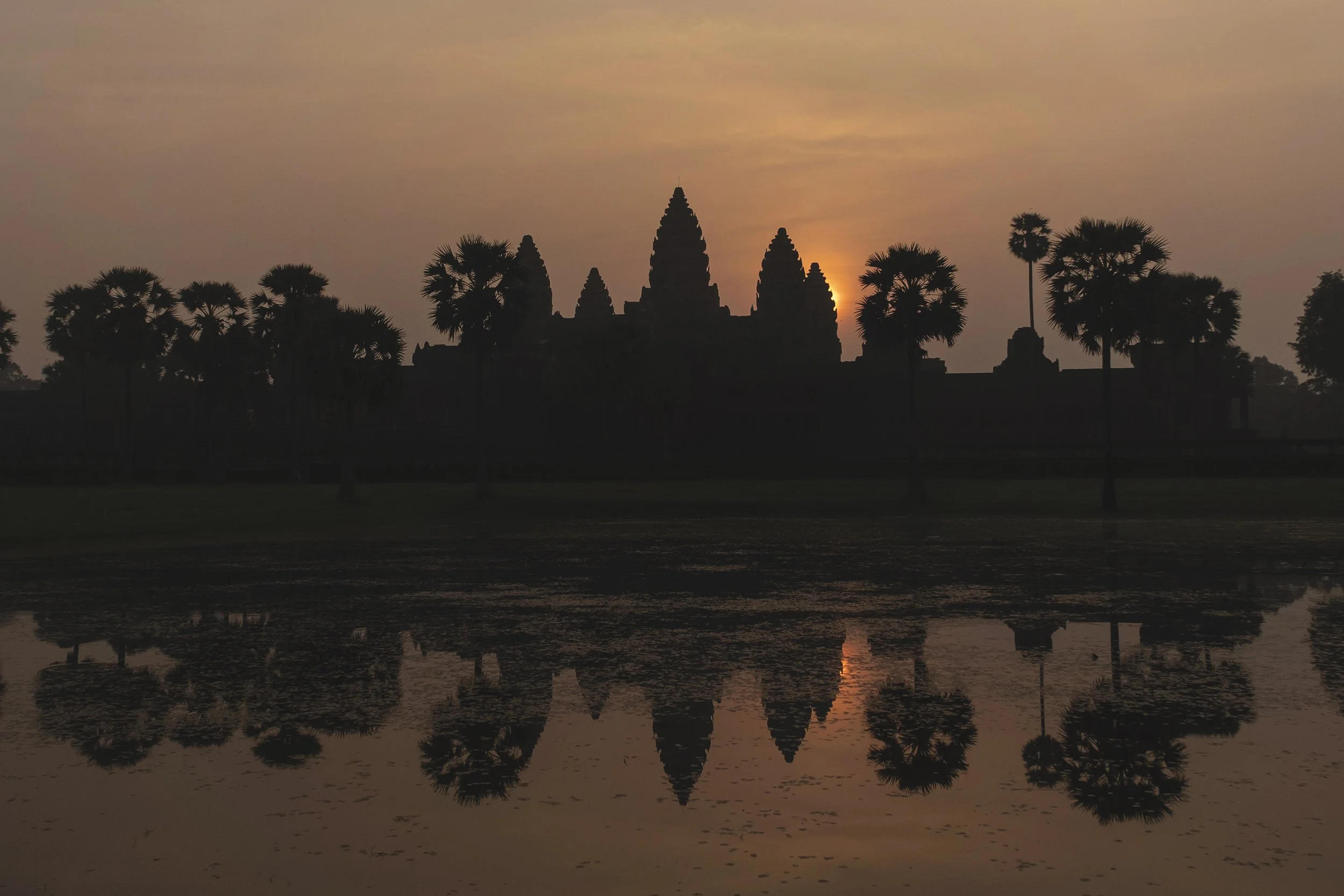 Silhouette du site d'Angkor Wat au lever du soleil, avec des palmiers et leur reflet dans l'eau calme.