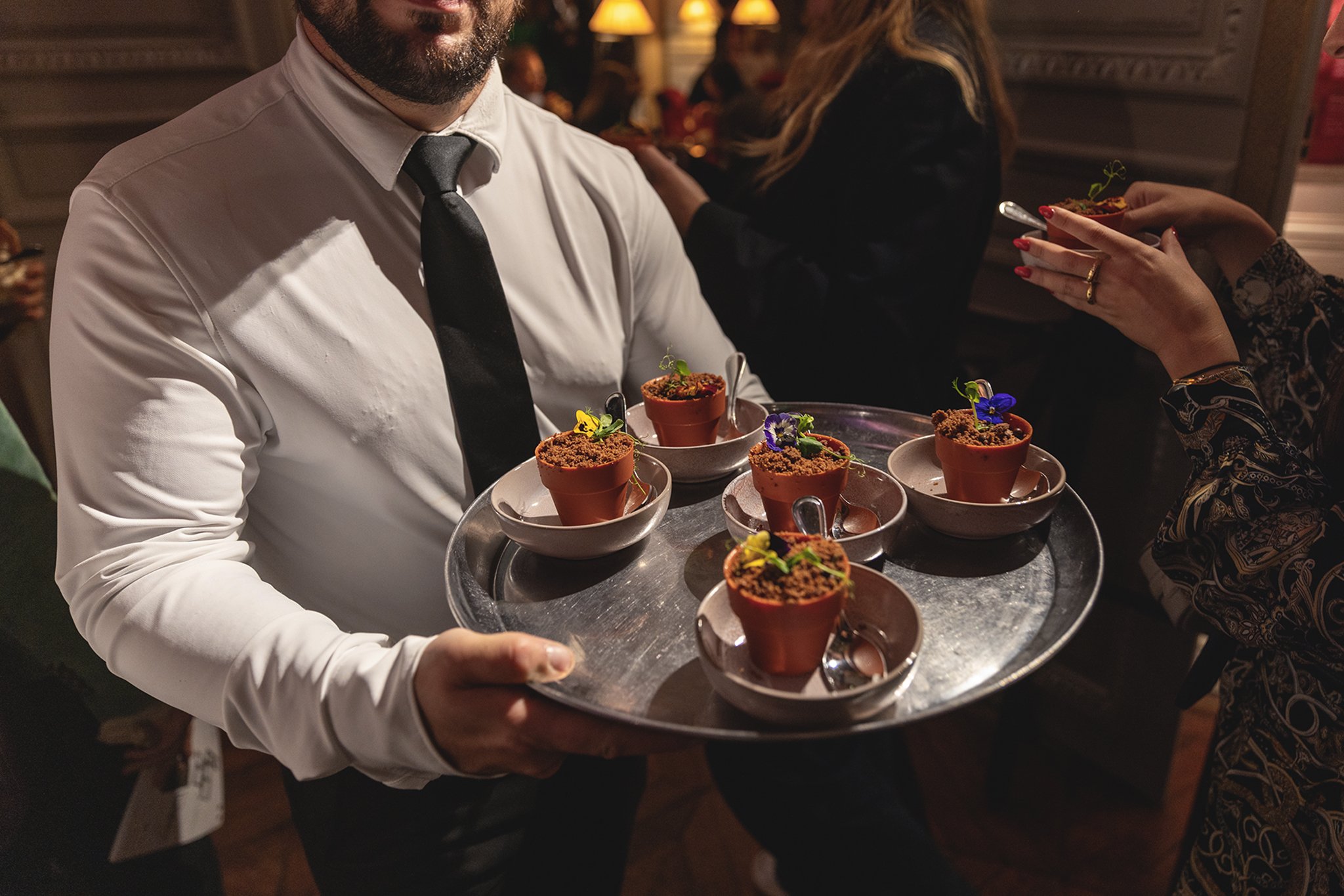 Un serveur tenant un plateau avec plusieurs desserts en petits pots en terre cuite, décorés de fleurs, lors d'un repas dans un restaurant.