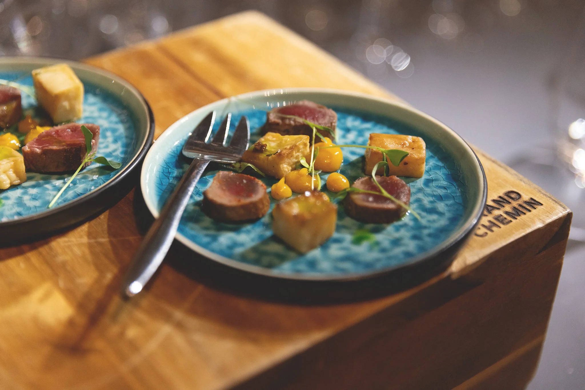 Une assiette de petite portion de viande rouge, de pommes de terre, de sauce jaune et de jeunes pousses sur une table en bois.
