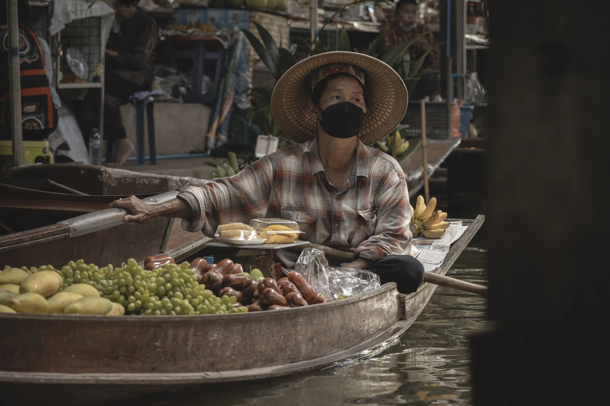 Femme sur un bateau-au marché flottant, vendant des fruits comme des raisins, des papayes, des bananes, portant un grand chapeau en paille et un masque noir.