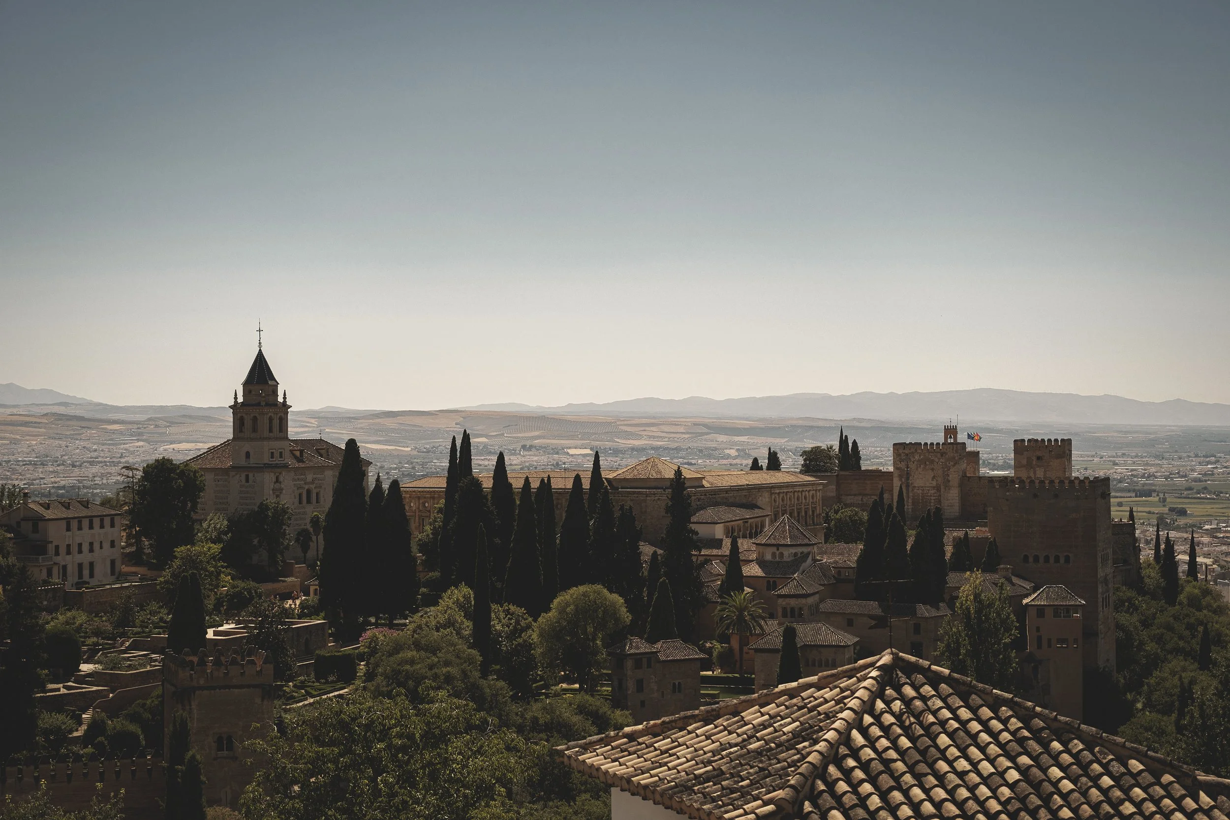 Vue d'un vieux centre-ville avec des bâtiments historiques en pierre, des tours et des toits en tuiles, entouré de végétation et de collines au loin sous un ciel clair.
