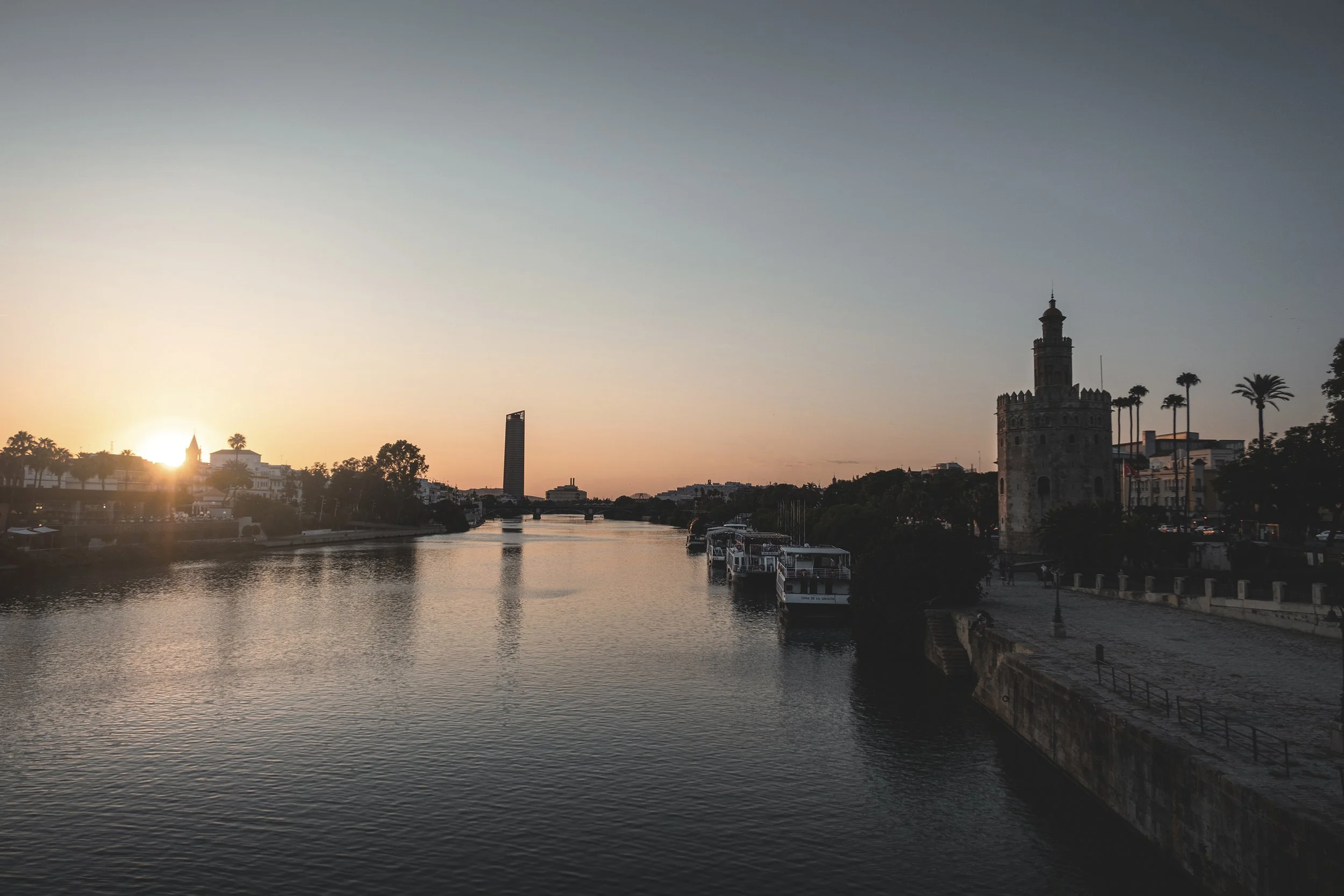 Coucher de soleil sur une rivière avec des bâtiments historiques et modernes, notamment une tour médiévale, des palmiers et des bateaux amarrés.