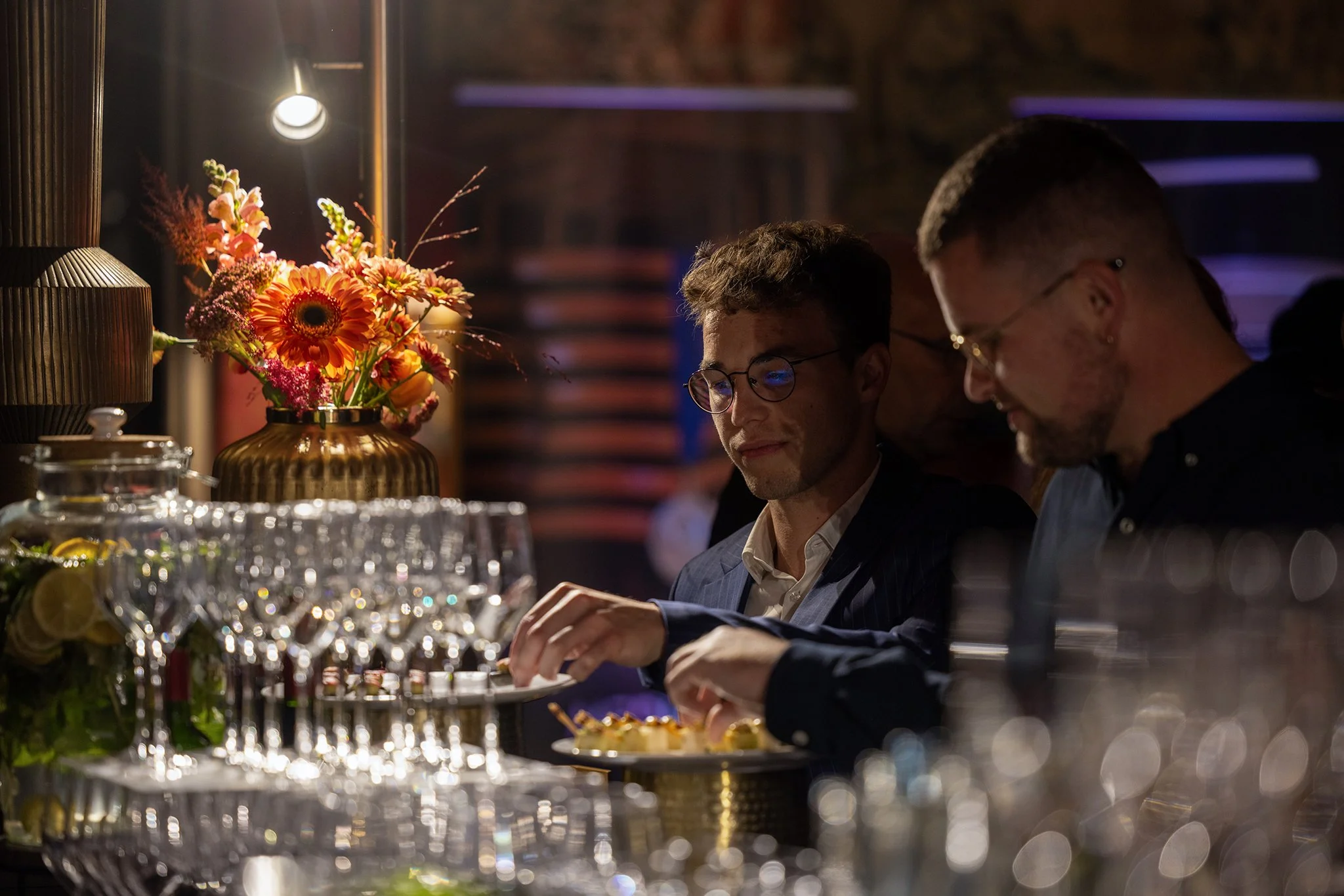 Un groupe de personnes assises autour d'une table lors d'un dîner, avec une vase remplie de fleurs colorées à gauche, sous un éclairage tamisé.