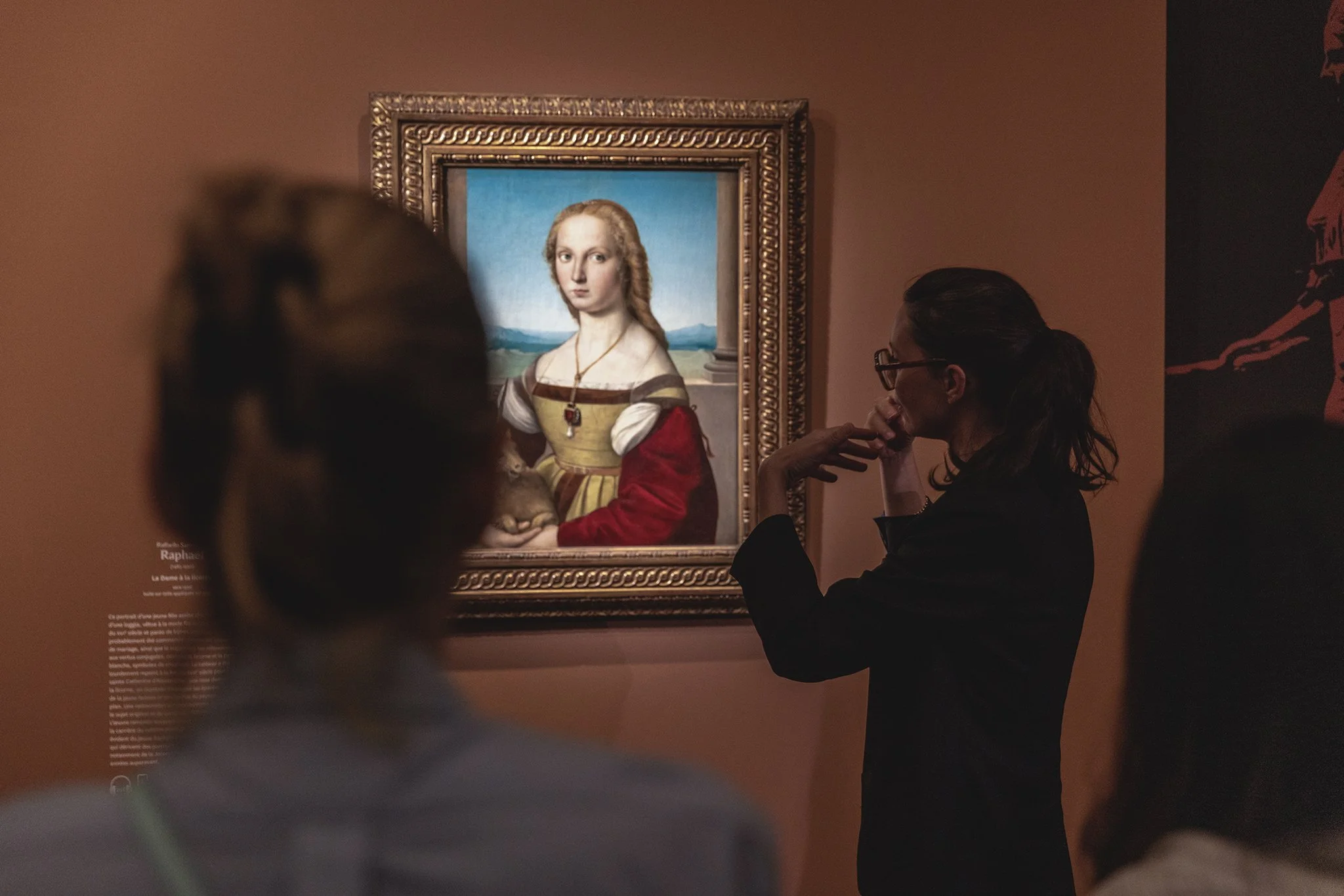 Une femme en lunettes observe une peinture de la Joconde dans un musée, avec une autre personne floue en premier plan.