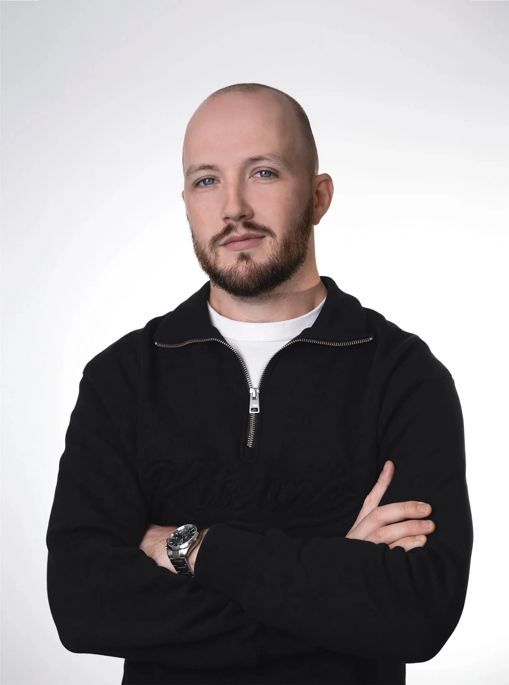 Homme souriant avec une barbe, portant une veste noire avec un zip, bras croisés, fond blanc