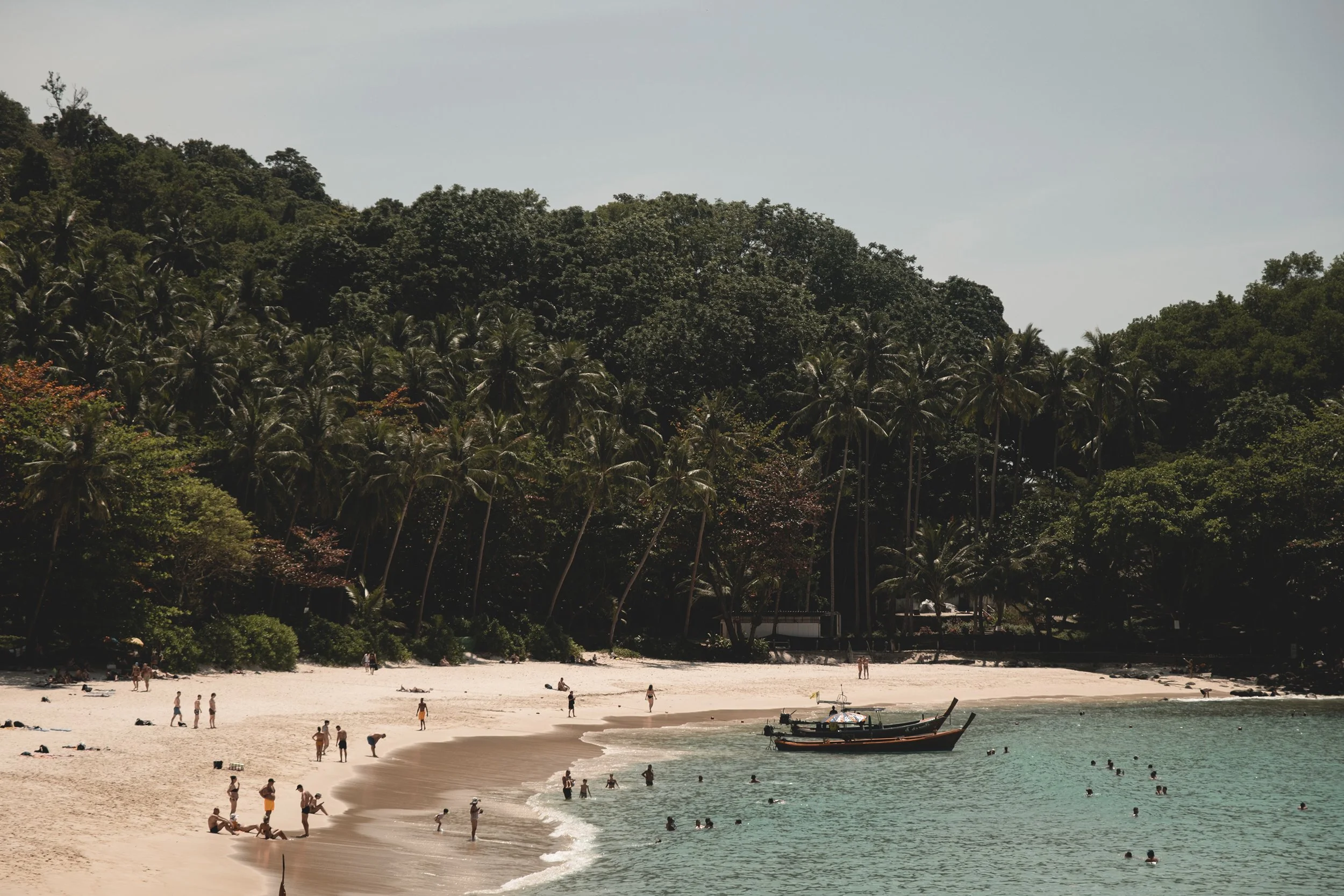 Plage avec des personnes nageant dans la mer, un bateau en bois et une forêt dense en arrière-plan