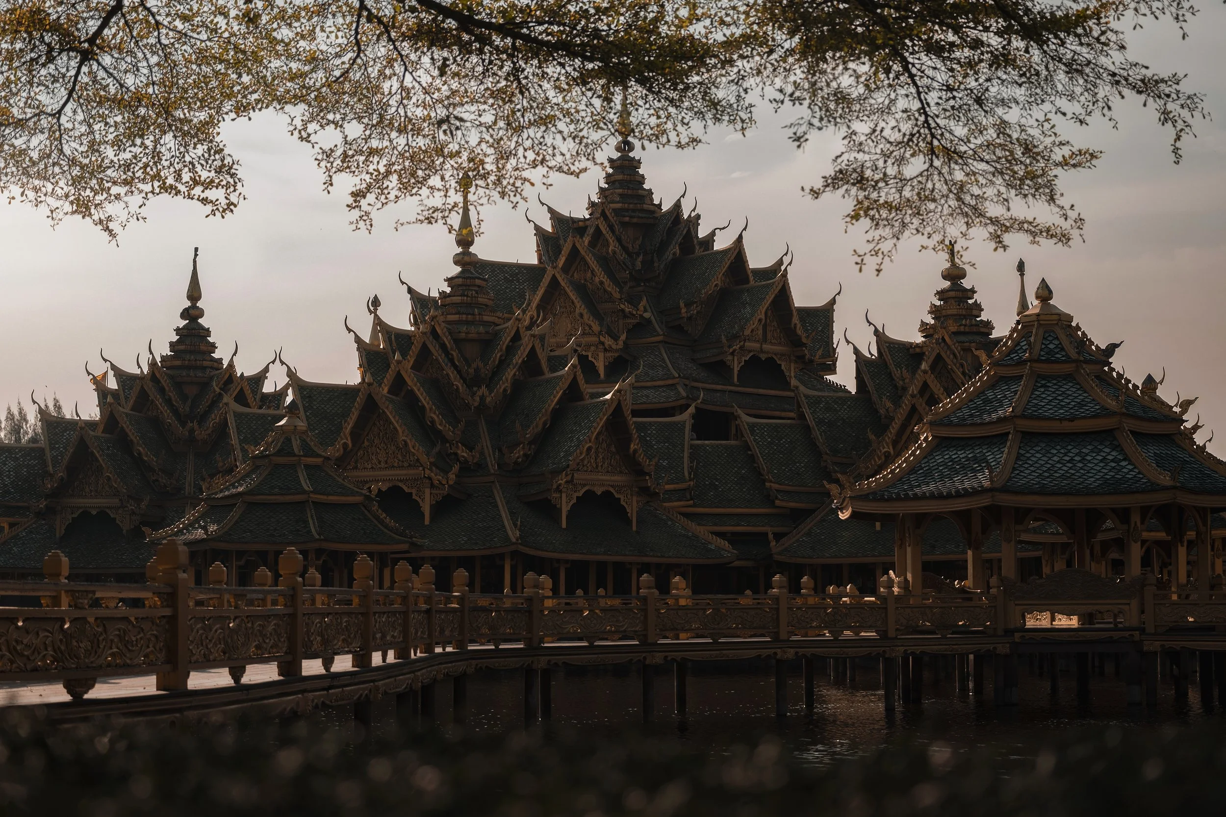 Vue nocturne d'un temple traditionnel thaïlandais en bois avec plusieurs toits superposés, situé au bord d'un lac, avec des arbres en arrière-plan.