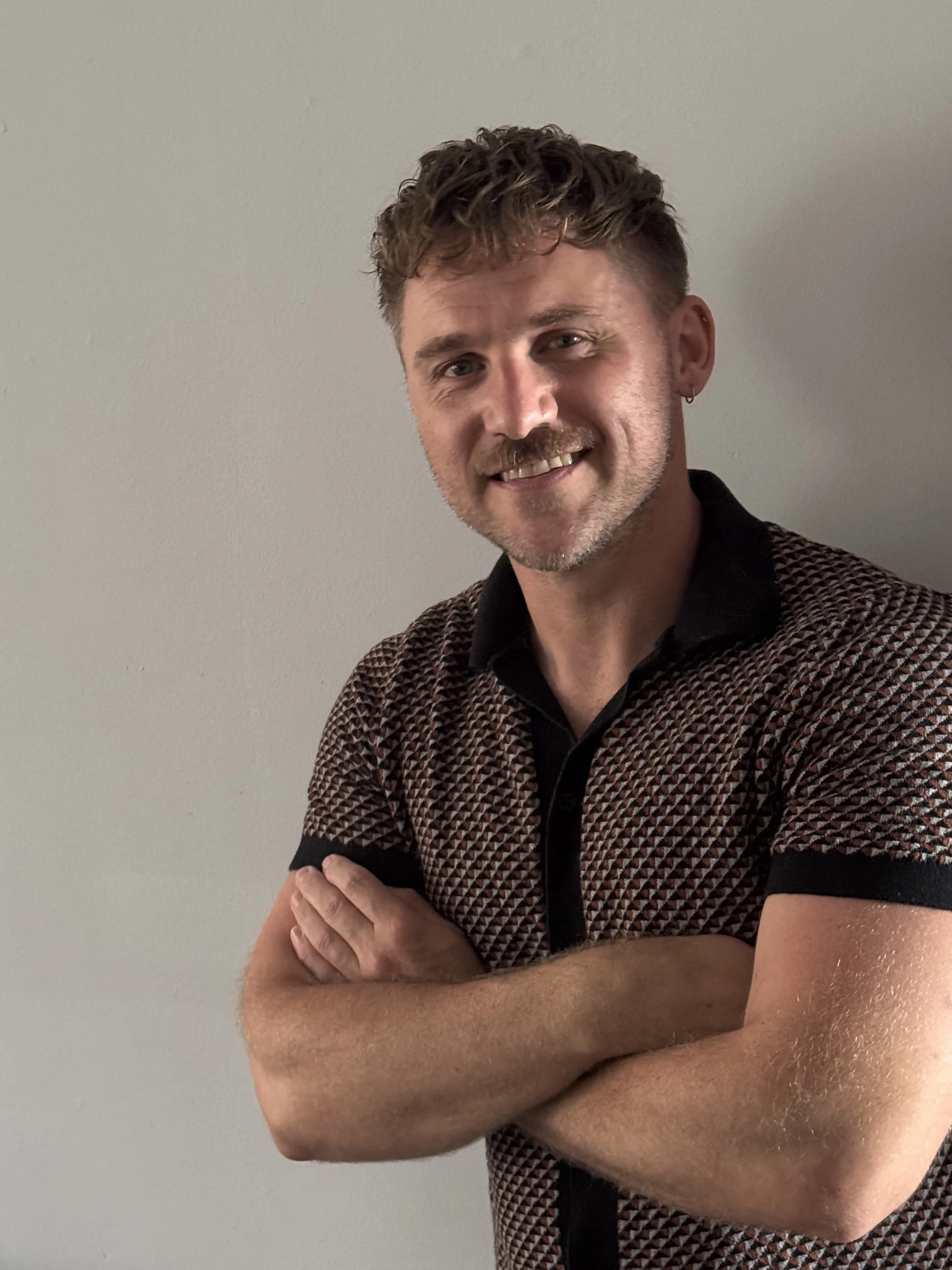 A man with short, curly brown hair and a beard, smiling with arms crossed, wearing a patterned short-sleeve shirt, standing against a plain white wall.