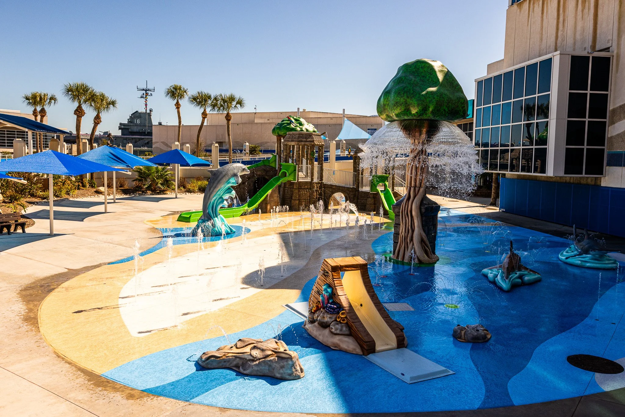 Empty children's water play area with dolphin and sea monster sculptures, slides, water features, and palm trees, outdoors in sunny weather.