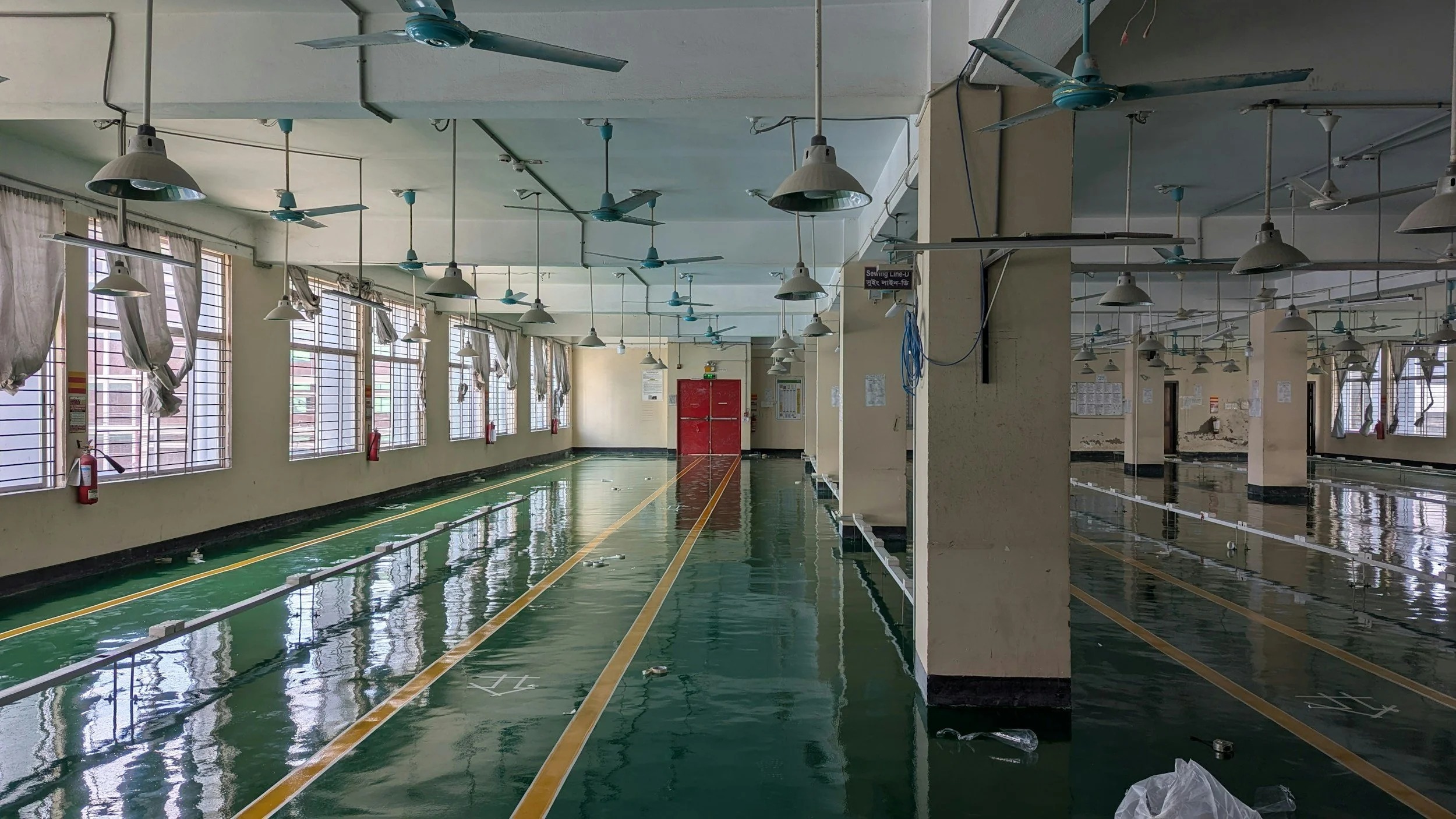 Empty indoor swimming pool or aquatic center with green painted floor, lined with yellow and white markings, and long windows with curtains on one side. Ceiling fans and hanging light fixtures are visible, and fire extinguishers are mounted by the windows. The pool appears to be under maintenance or cleaning.