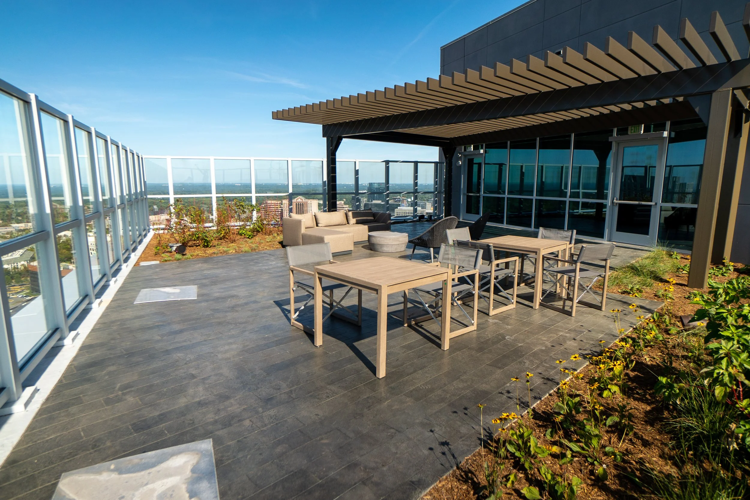 Rooftop patio with outdoor furniture, including tables, chairs, and a seating area, enclosed with glass railings, with cityscape in the background under a clear blue sky.
