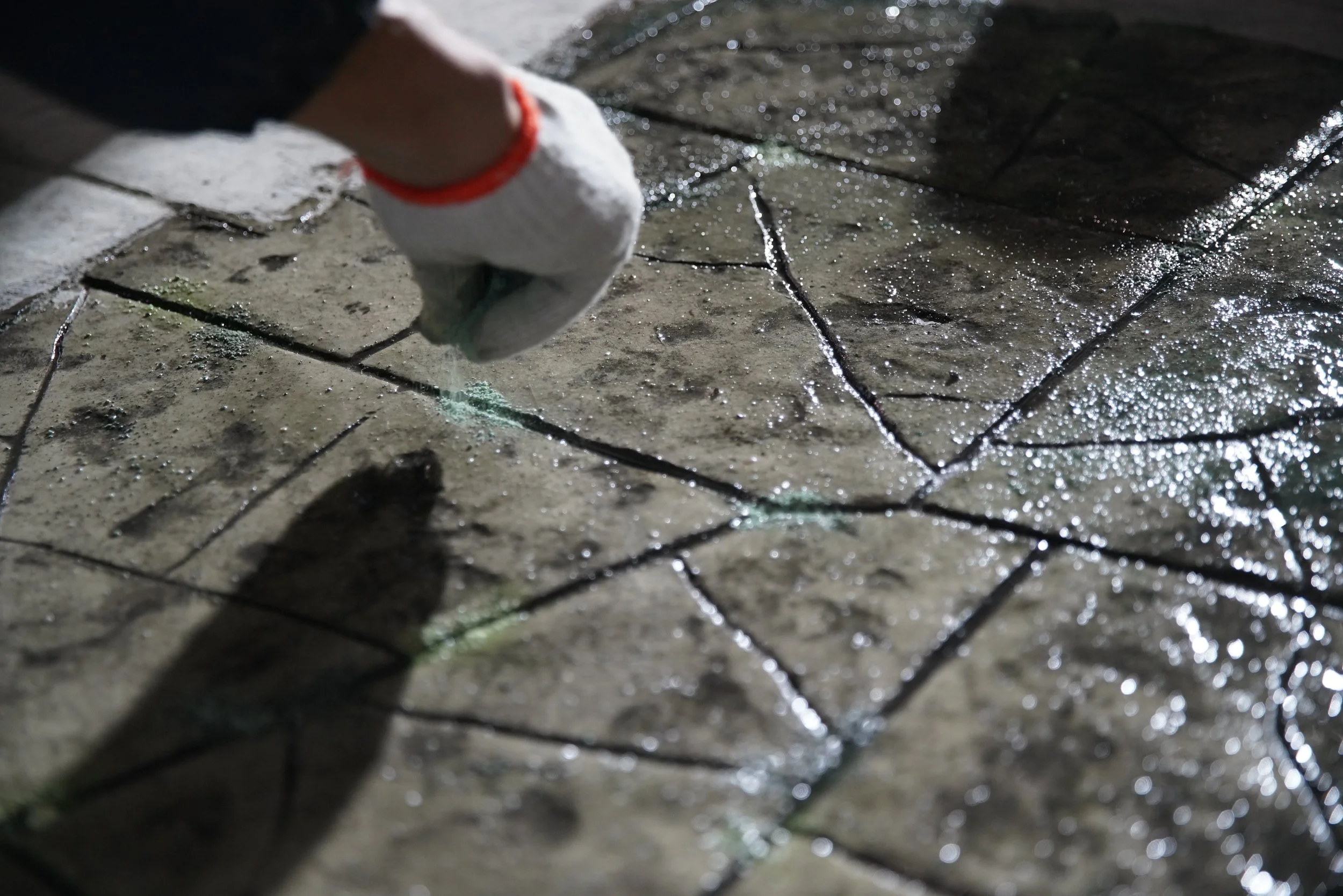 Close-up of a person's hand wearing a white glove with a red trim, applying grout to the cracks of wet, stained concrete paving stones.