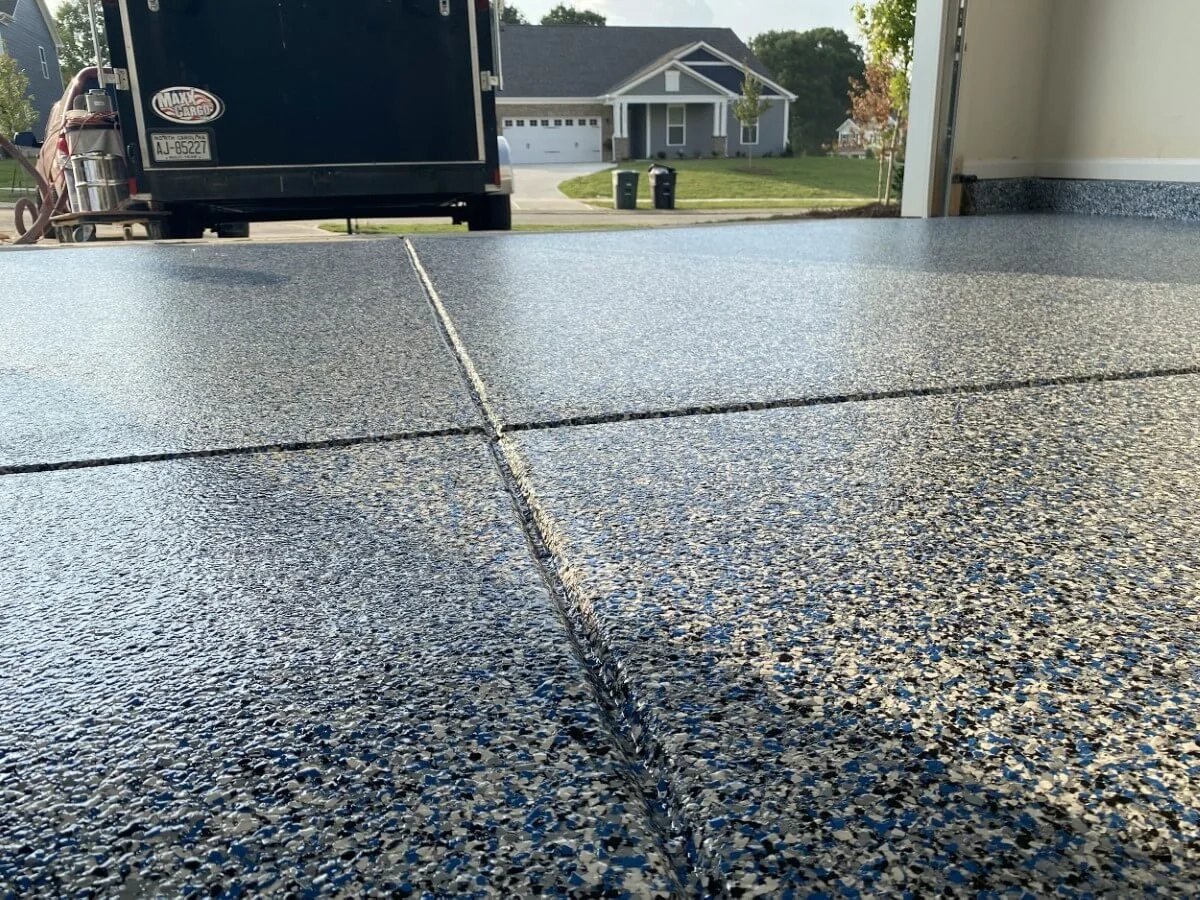 Close-up of a garage floor with freshly sealed black and gray patterned tiles, with a suburban neighborhood, house, and trash bins visible outside.