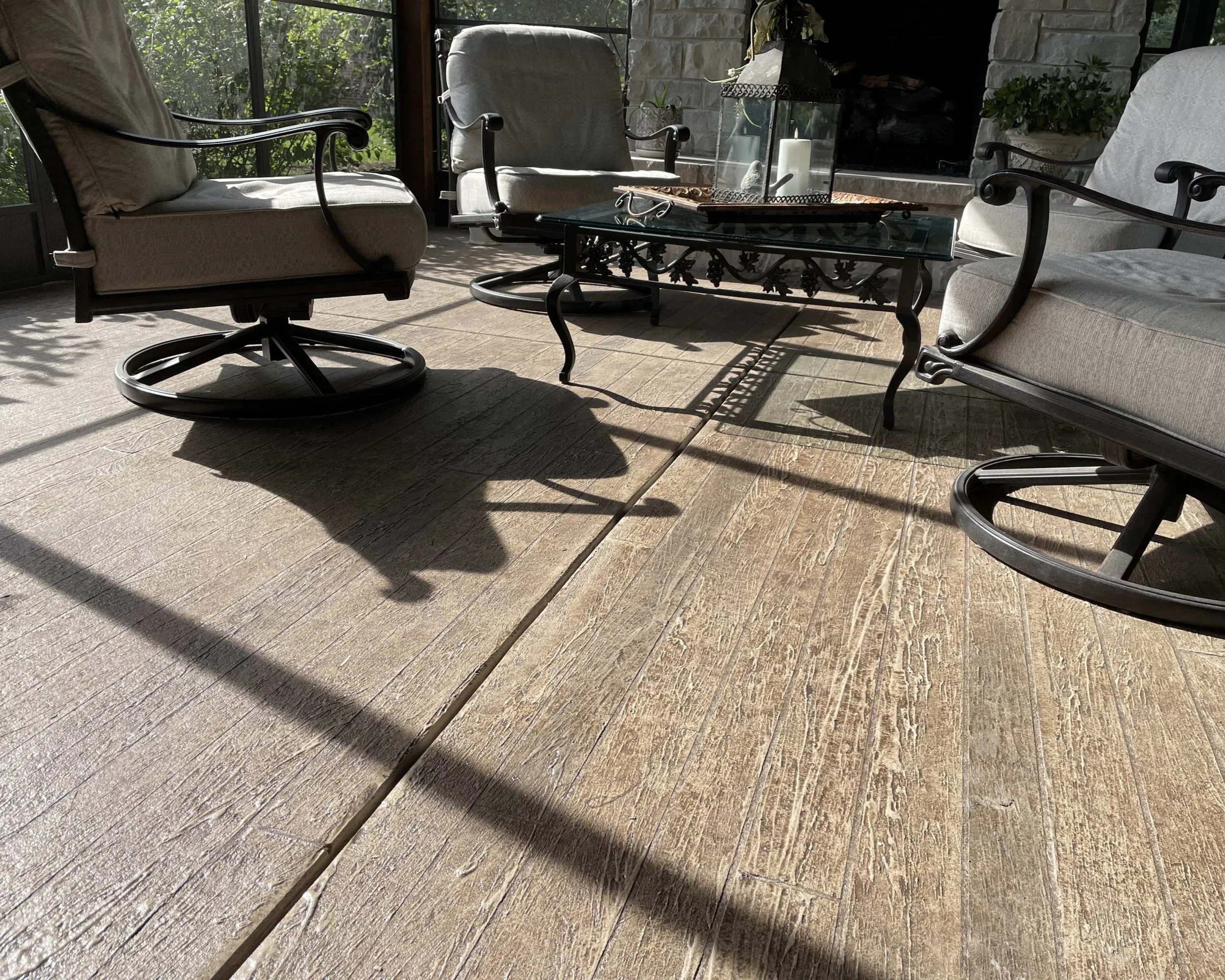 Enclosed sunroom with four cushioned chairs, a glass-topped ornate black metal table, and potted plants near a stone fireplace, illuminated by natural sunlight.