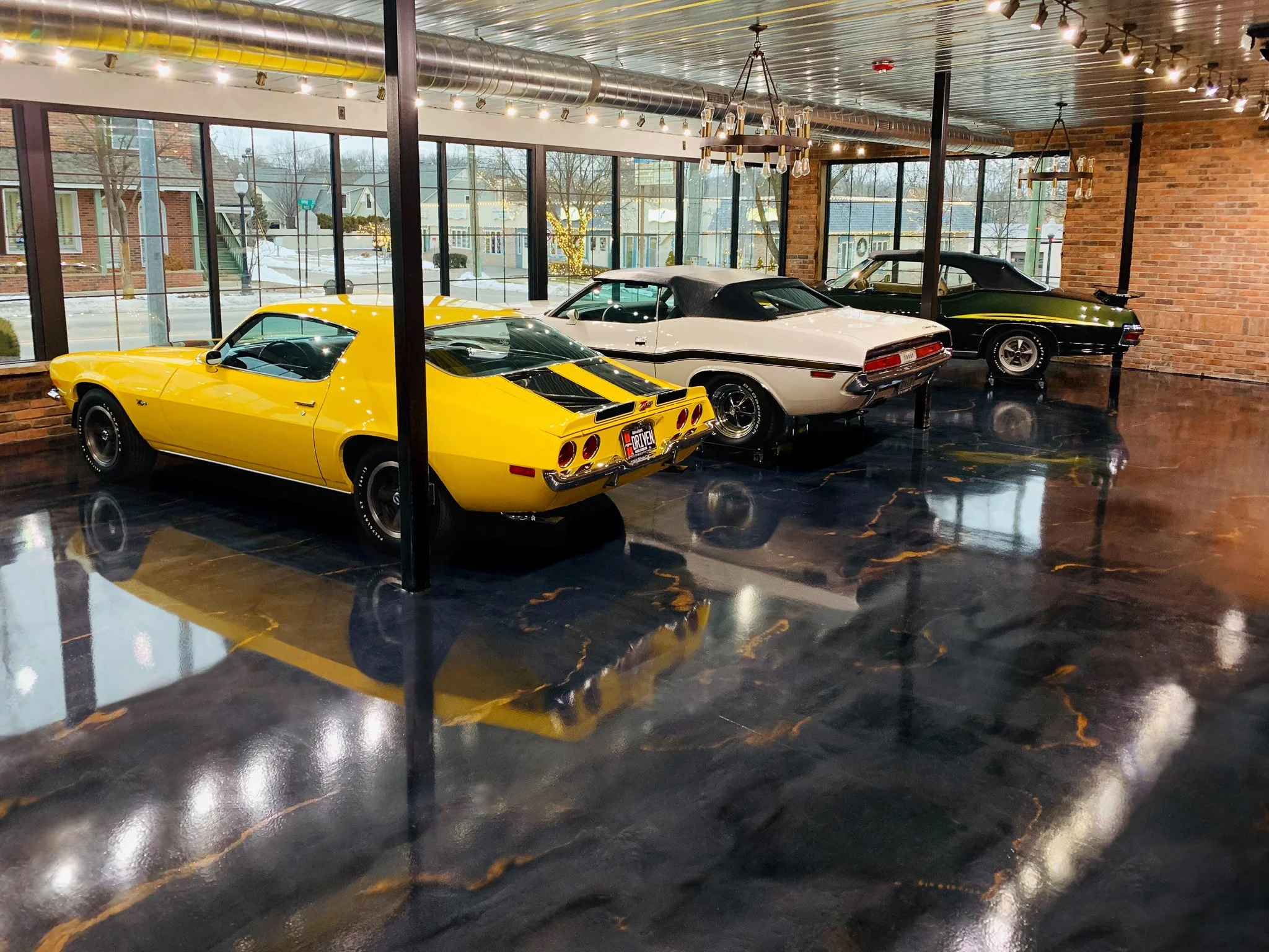Three classic cars are displayed inside a showroom with large windows and brick walls, including a yellow Chevrolet Corvette, a white convertible, and a black muscle car.