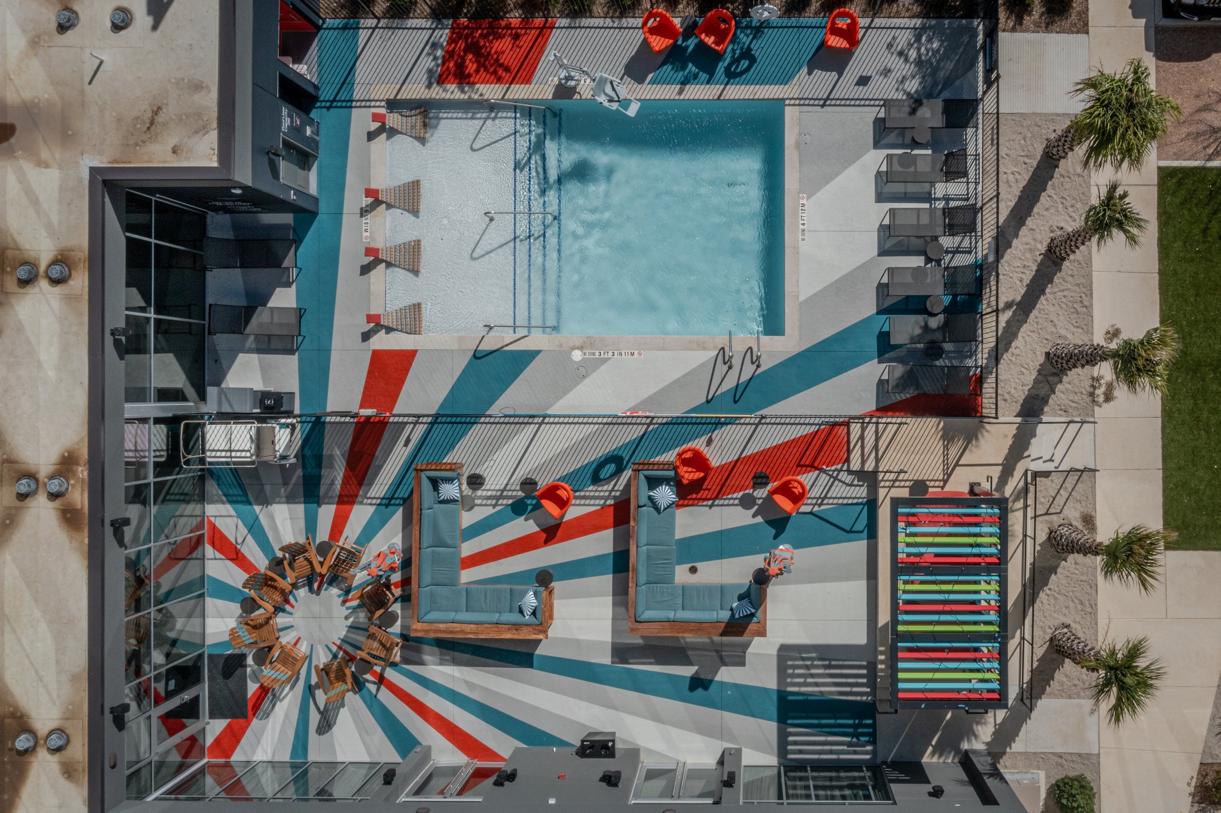 Aerial view of a swimming pool area with modern lounge chairs, orange slides, colorful benches, and decorative palm trees.
