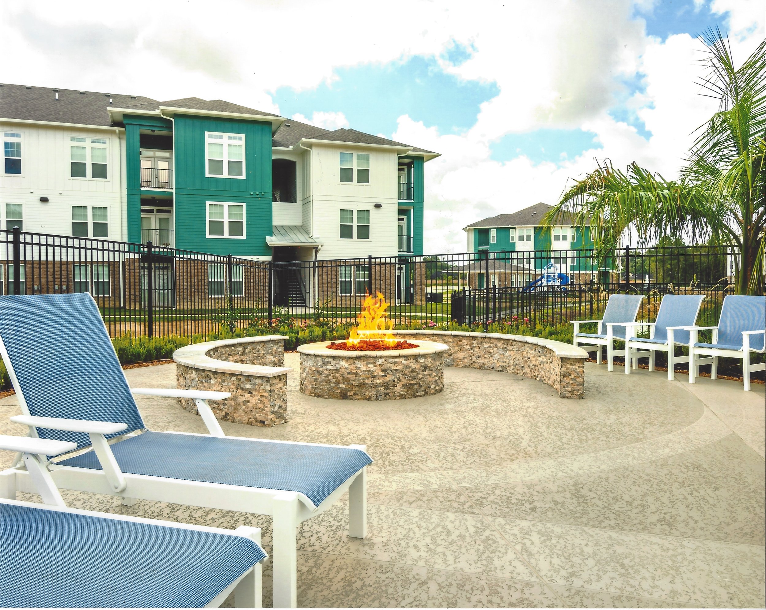 A courtyard with a fire pit, surrounded by blue chairs and a black metal fence, with residential buildings in the background and palm trees to the right.