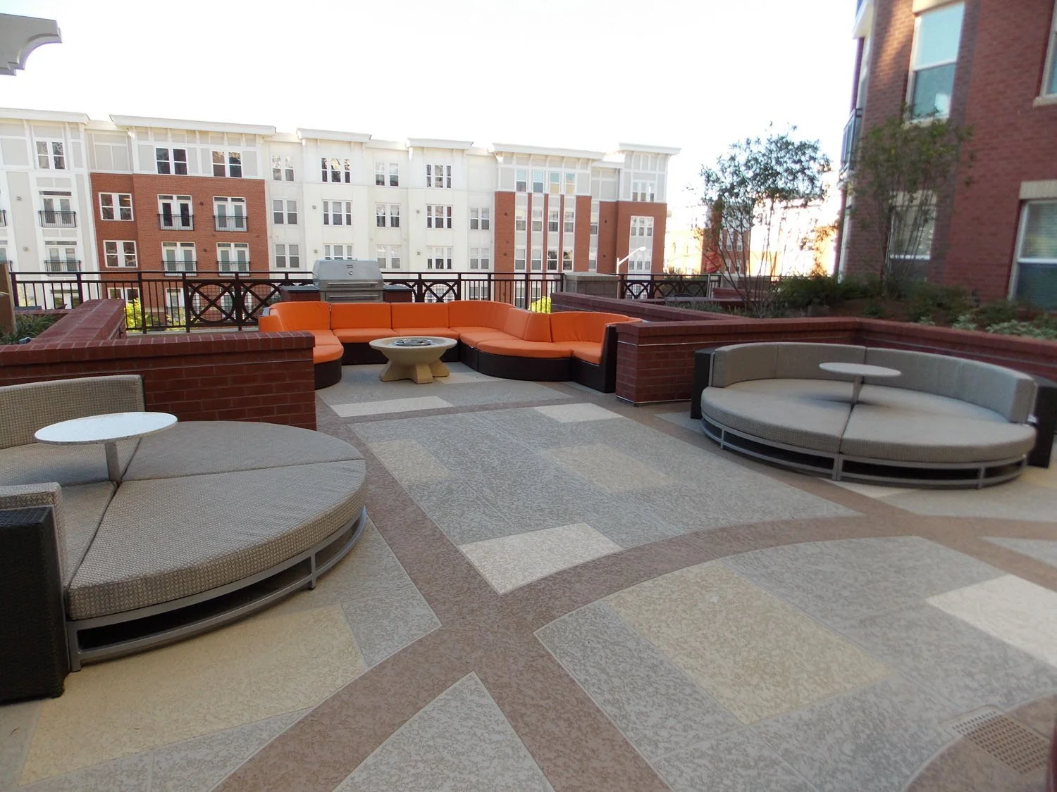 Outdoor patio area with curved seating, orange and beige cushions, tables, a barbecue grill, and a cityscape backdrop with residential buildings.