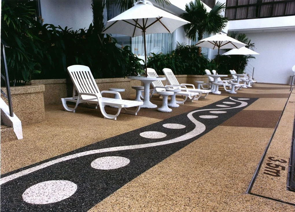 Poolside loungers with white umbrellas, tropical plants, and a decorative winding black and white line on the ground near a building with glass windows.