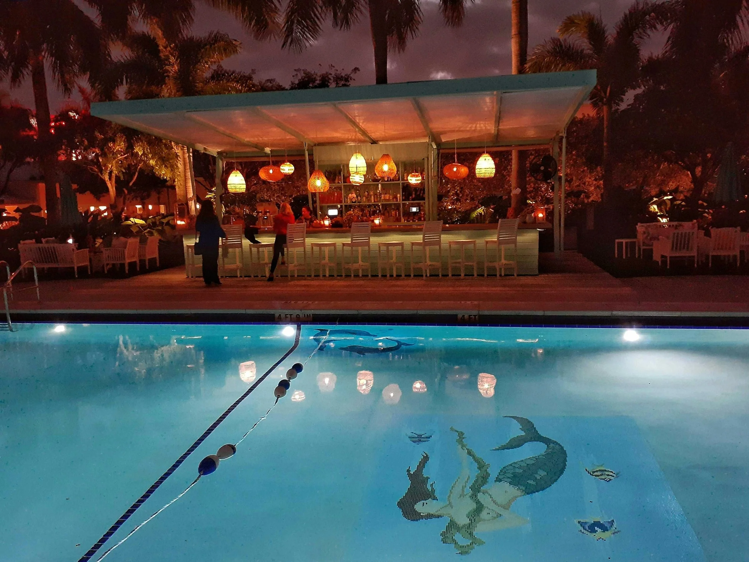 Nighttime view of a pool with a mosaic of a mermaid and fish, illuminated by underwater lights. In the background, inside a bar area, are hanging lamps and bartenders serving drinks, with palm trees and lounge chairs around the pool area.