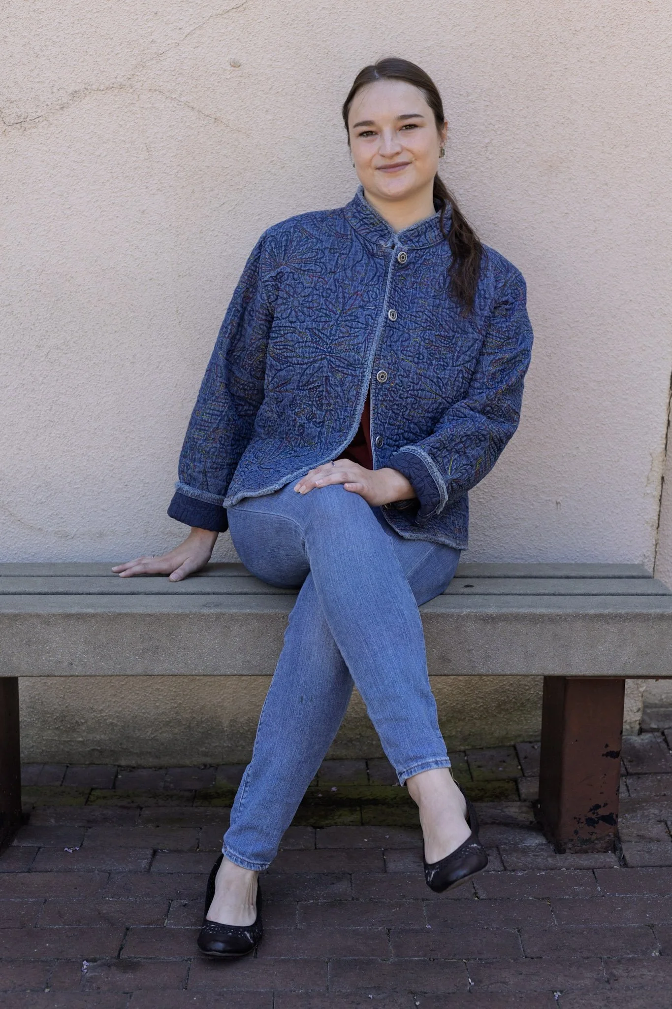A woman sitting on a wooden bench against a beige wall, wearing a blue patterned jacket, jeans, and black shoes, with her hair tied back.