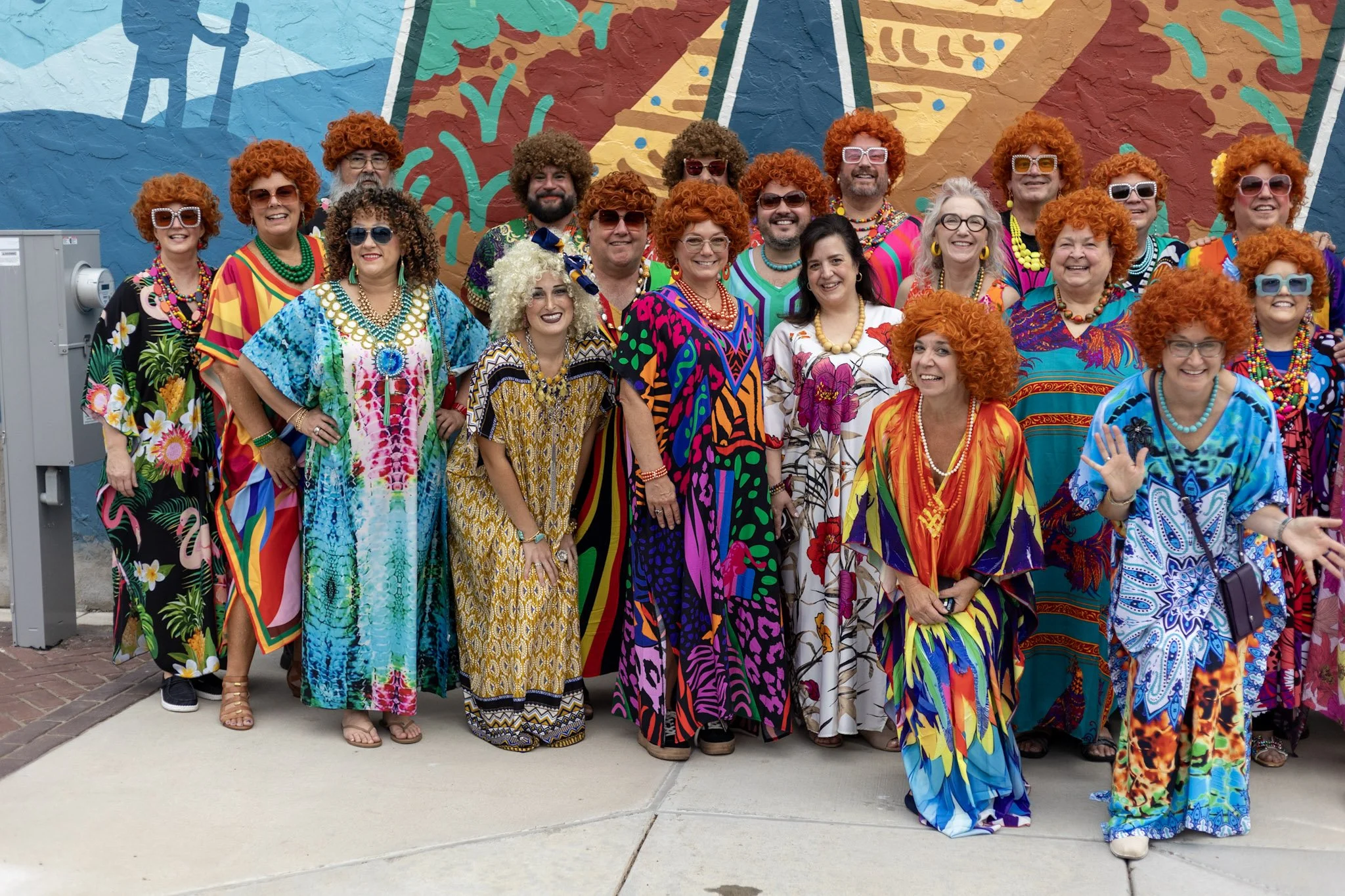 Group of women and men dressed in colorful, retro 70s style clothing, wearing curly red wigs and sunglasses, posing in front of a vibrant, abstract mural.