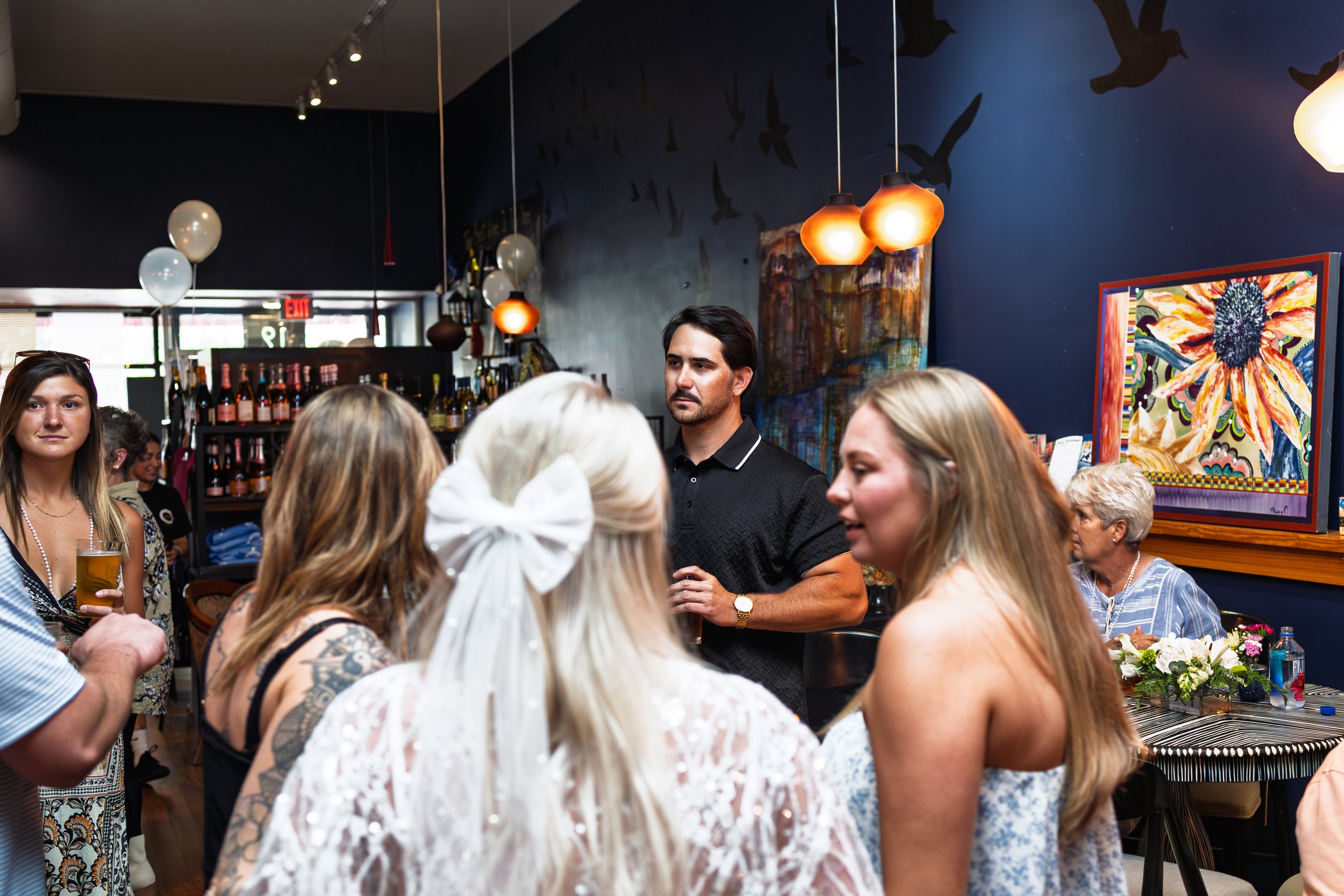A group of people socializing at an art gallery or restaurant with paintings on the wall and hanging lights.