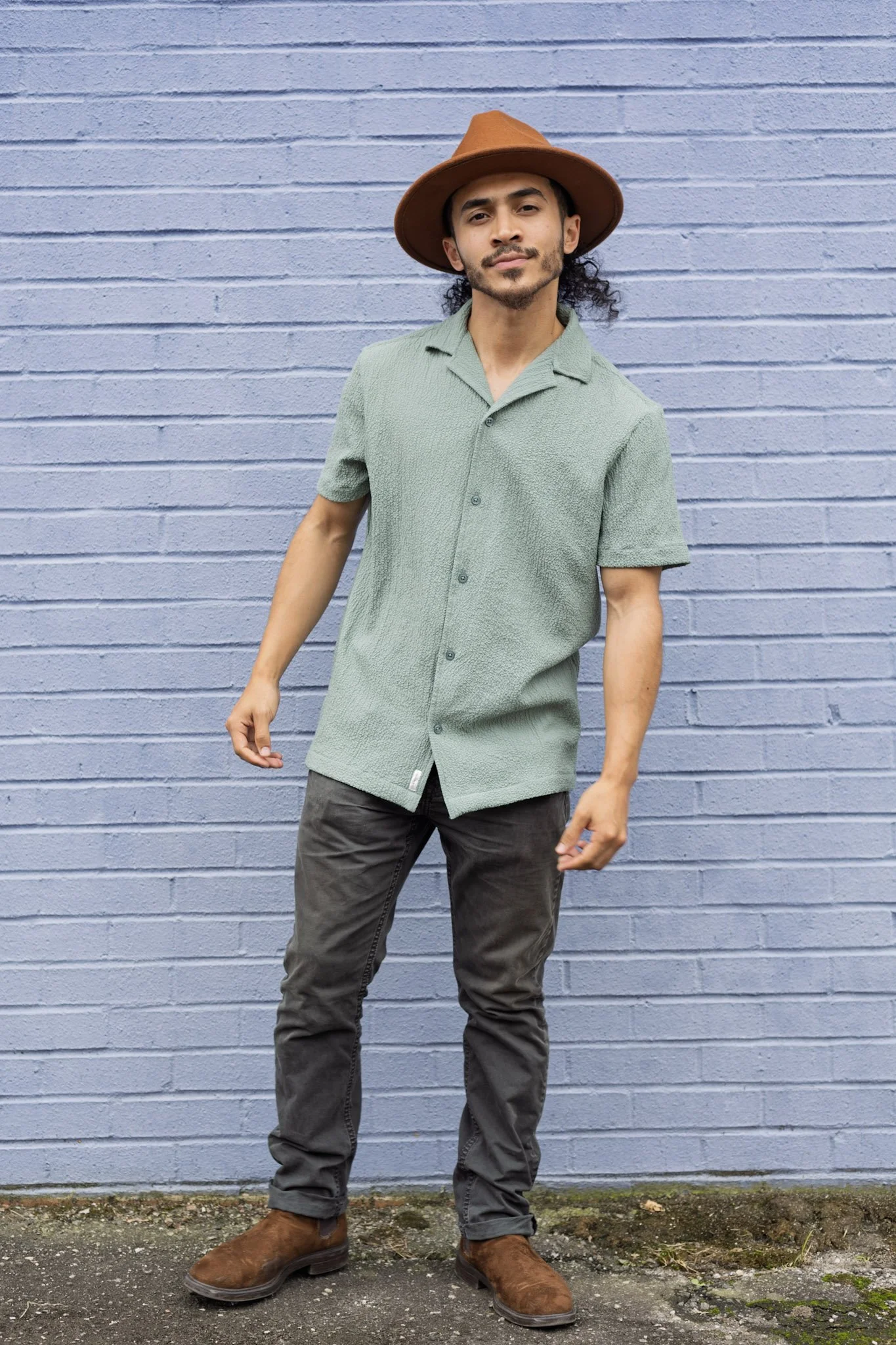 A man wearing a wide-brimmed brown hat, a light green short-sleeved textured shirt, dark gray pants, and brown boots, standing in front of a purple brick wall.