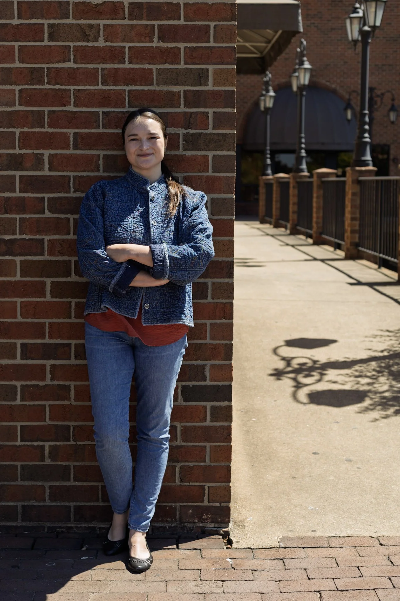 A woman standing outdoors against a brick wall, with arms crossed, wearing a blue jacket, red shirt, blue jeans, and black shoes. Shadows of street lamps are visible on the sidewalk.