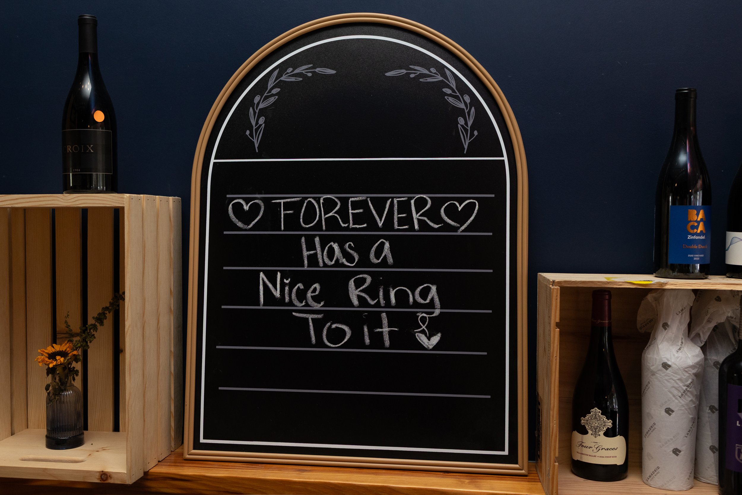 A blackboard sign with white chalk writing that says 'Forever has a Nice Ring To It' with hearts, surrounded by bottles of wine and wooden shelves.