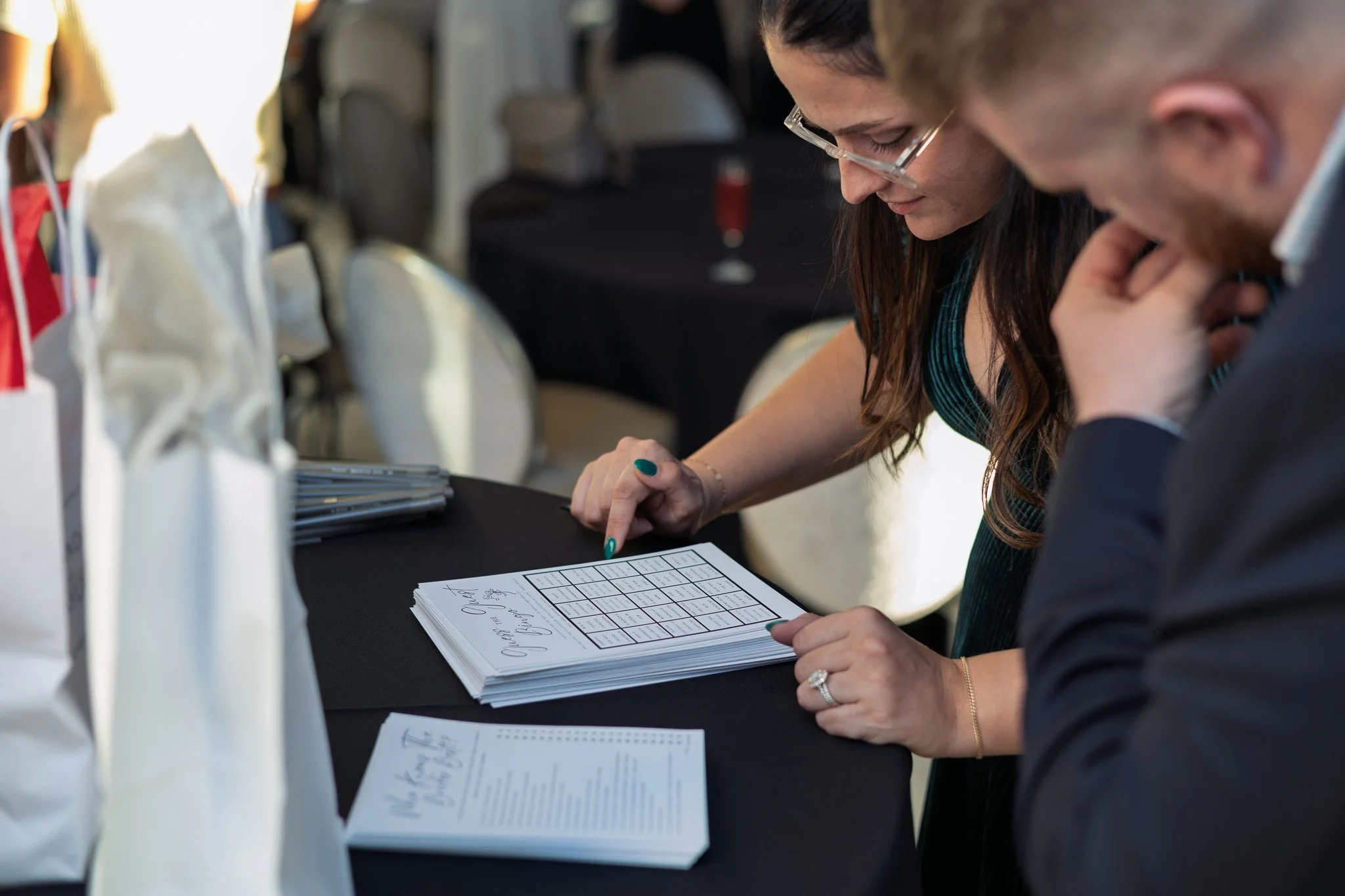 A woman and a man look at a sheet of paper with a grid on it, possibly at a conference or event, with handbags on a table and chairs in the background.