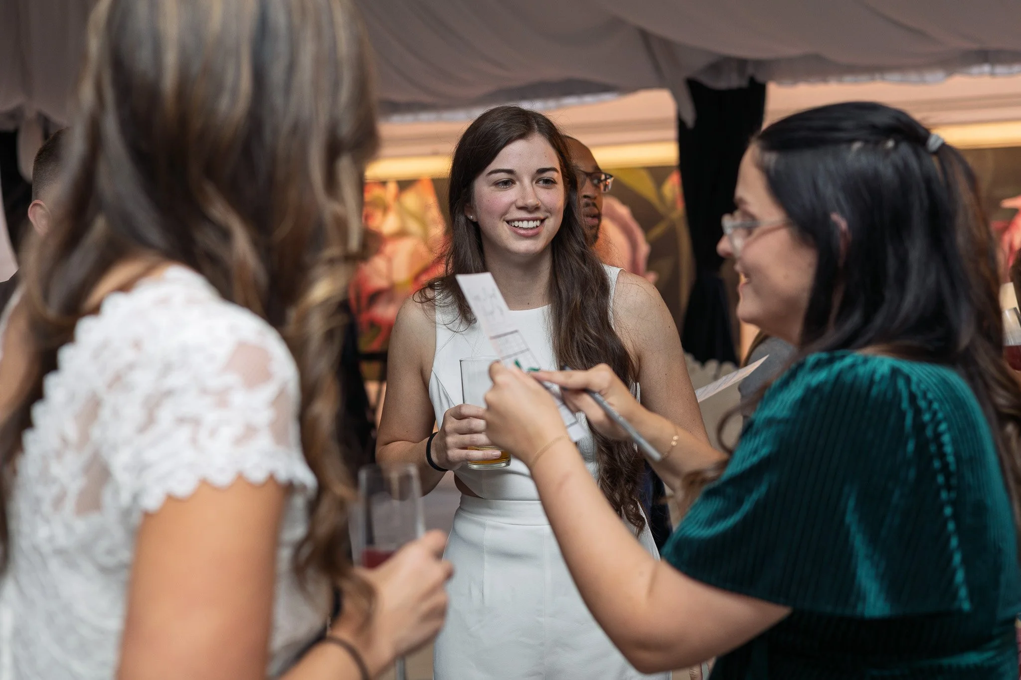 Young woman with long brown hair wearing a white dress holding a drink, smiling and talking with two women at a social event indoors.